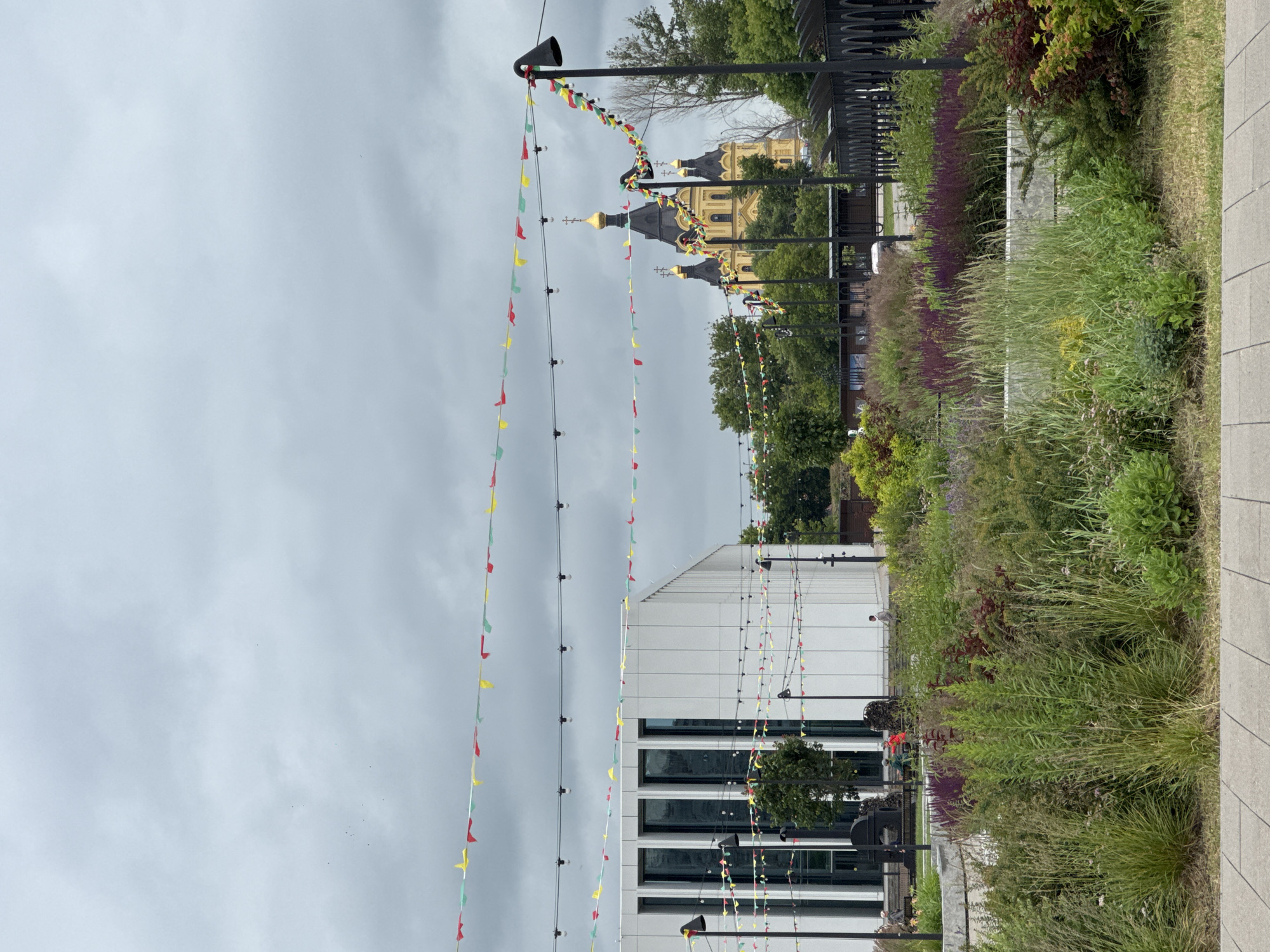 The image shows a view of the Nizhny Novgorod Kremlin's Alexander Nevsky Cathedral, a prominent yellow and black domed structure, seen from a landscaped area. In the foreground, there is a lush garden with various plants and flowers in shades of green and purple. String lights and colorful triangular flags are strung across the scene, suggesting a festive atmosphere or an event. To the left, a modern white building with large windows is visible. The sky is overcast, indicating cloudy weather. Several indistinct figures can be seen in the distance, some sitting on benches near the modern building. The overall scene depicts an urban park or public space in Nizhny Novgorod, Russia, with a blend of historical and modern architecture amidst natural landscaping.