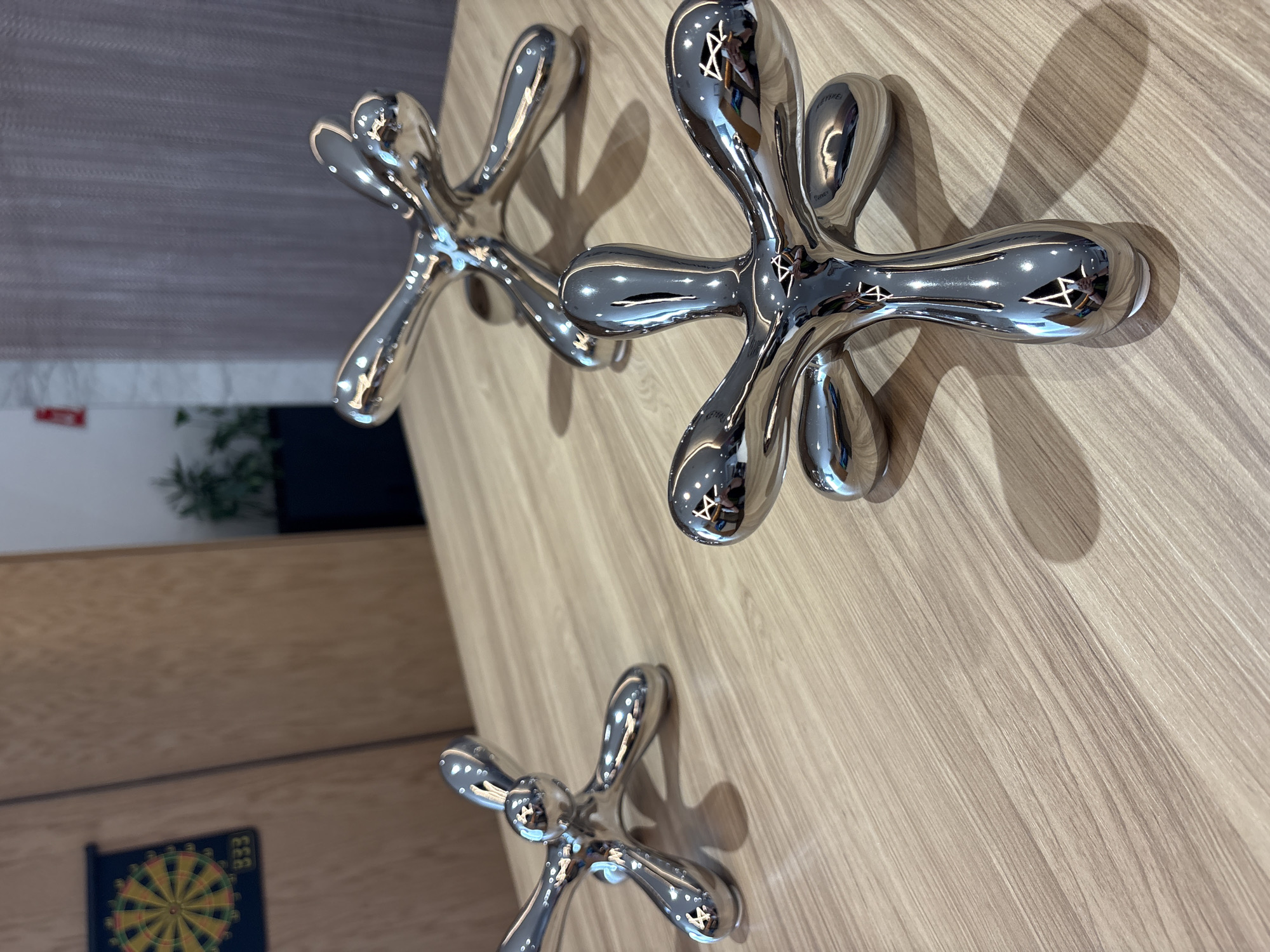The image shows a close-up view of a wooden surface with three metal, jack-shaped objects attached to it. The objects are shiny and reflect light, giving them a distorted, mirrored appearance. Their shadows are visible on the wood. In the background, partially out of focus, is a room interior with a glimpse of a dartboard and a plant. The setting appears to be inside a building. There is no information provided in the image to indicate the location in Mexico City, Mexico.