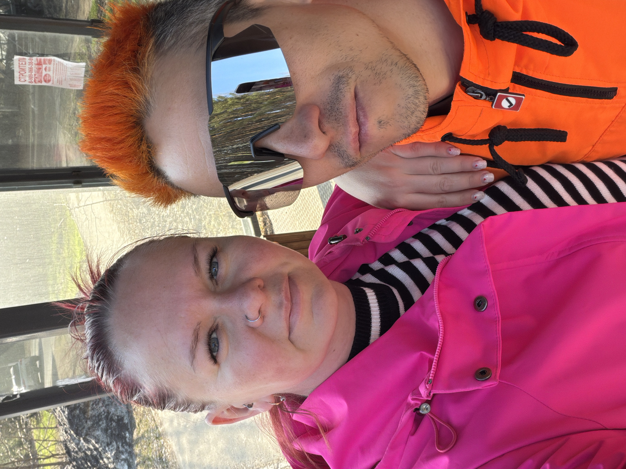 Two individuals pose for a selfie. One person has short, orange-colored hair and wears sunglasses that reflect an outdoor environment with trees and the sky. They are wearing an orange jacket with black accents and black drawstrings. The other person has short, reddish-brown hair and a nose ring. They are wearing a black and white striped sweater and a bright pink jacket. The background appears to be a window with a view of trees and foliage. A printed sticker can be seen on the window. The image does not provide definitive information to confirm the location is in Chaikovsky, Russia.