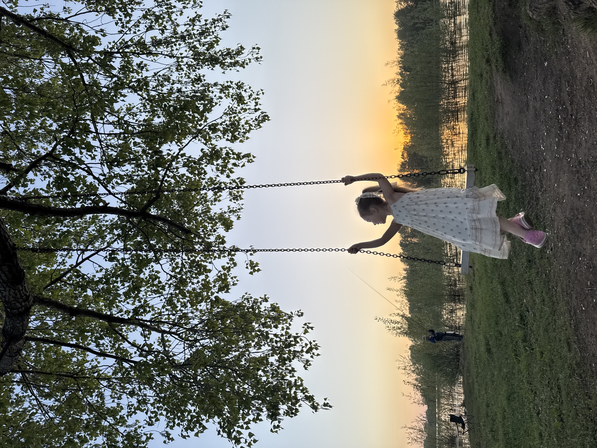 A young girl wearing a white dress and pink shoes is swinging on a swing set in Chaikovsky, Russia. The swing set is located near a body of water lined with trees and foliage. A large tree with green leaves is visible to the left of the swing set. Two people are standing near the water's edge. The sky is a gradient of colors suggesting either a sunrise or sunset.