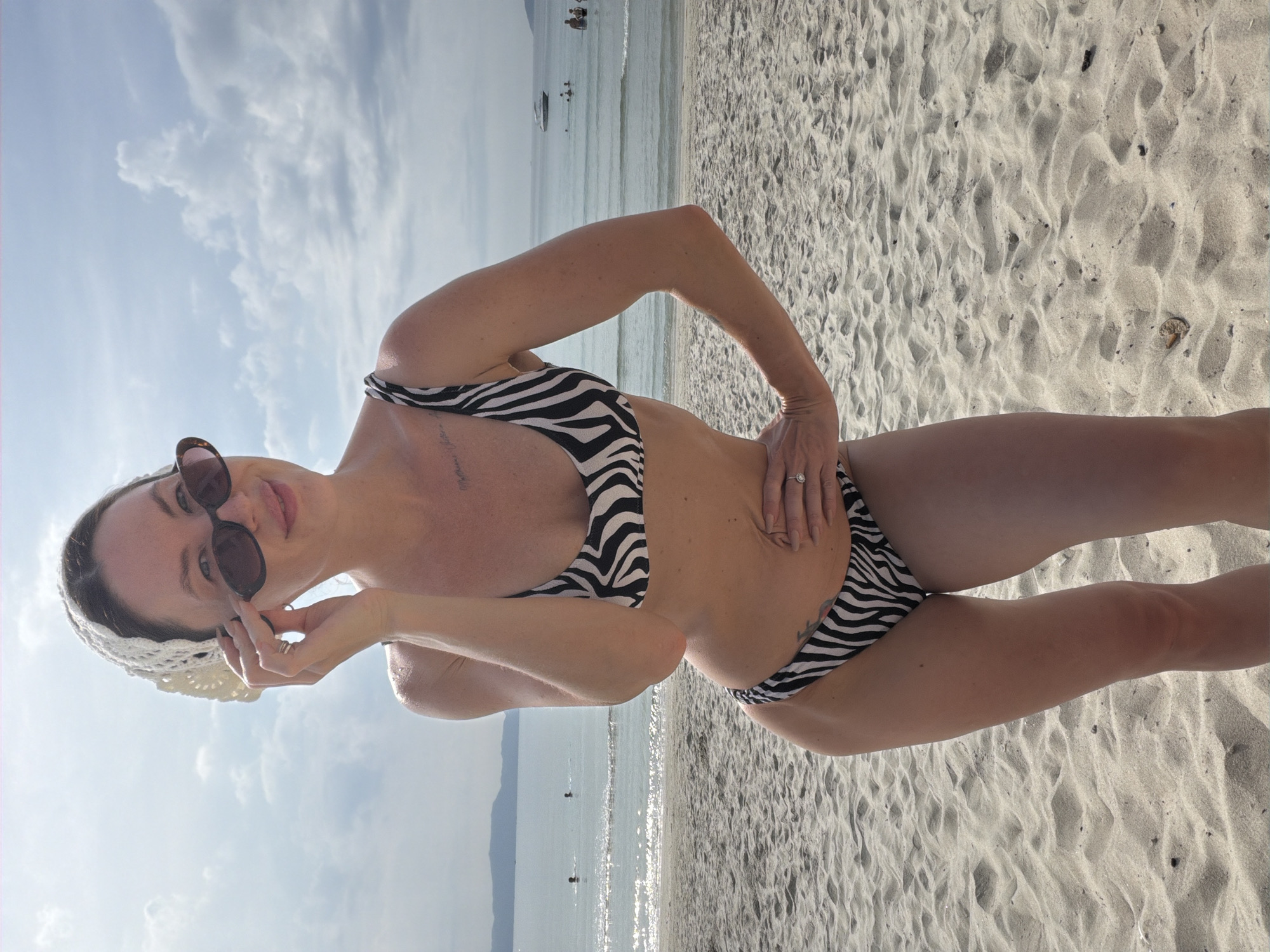 A woman in a zebra-print bikini and sunglasses poses on a sandy beach with the ocean and sky in the background. She is wearing a white crochet headscarf and has one hand on her hip and the other adjusting her sunglasses. The water has gentle waves and a few distant boats and people. The sand is light brown and textured with footprints. The overall lighting suggests a sunny day. The location is Dong Ninh Hoa Ward, Vietnam.