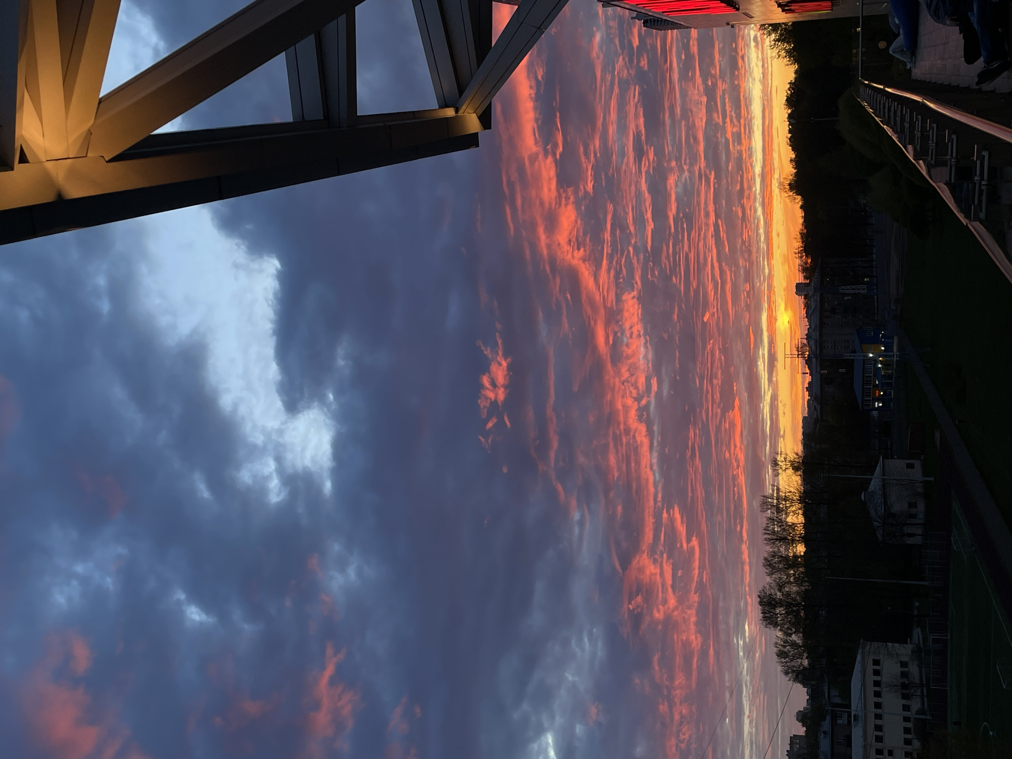 The image captures a sunset view in Saint Petersburg, Russia. The sky is filled with dramatic clouds tinged with orange and red hues from the setting sun. A building structure is partially visible in the foreground on the left side of the frame, and a railing and walkway are seen on the right. In the distance, there's a silhouette of buildings and trees against the bright horizon.