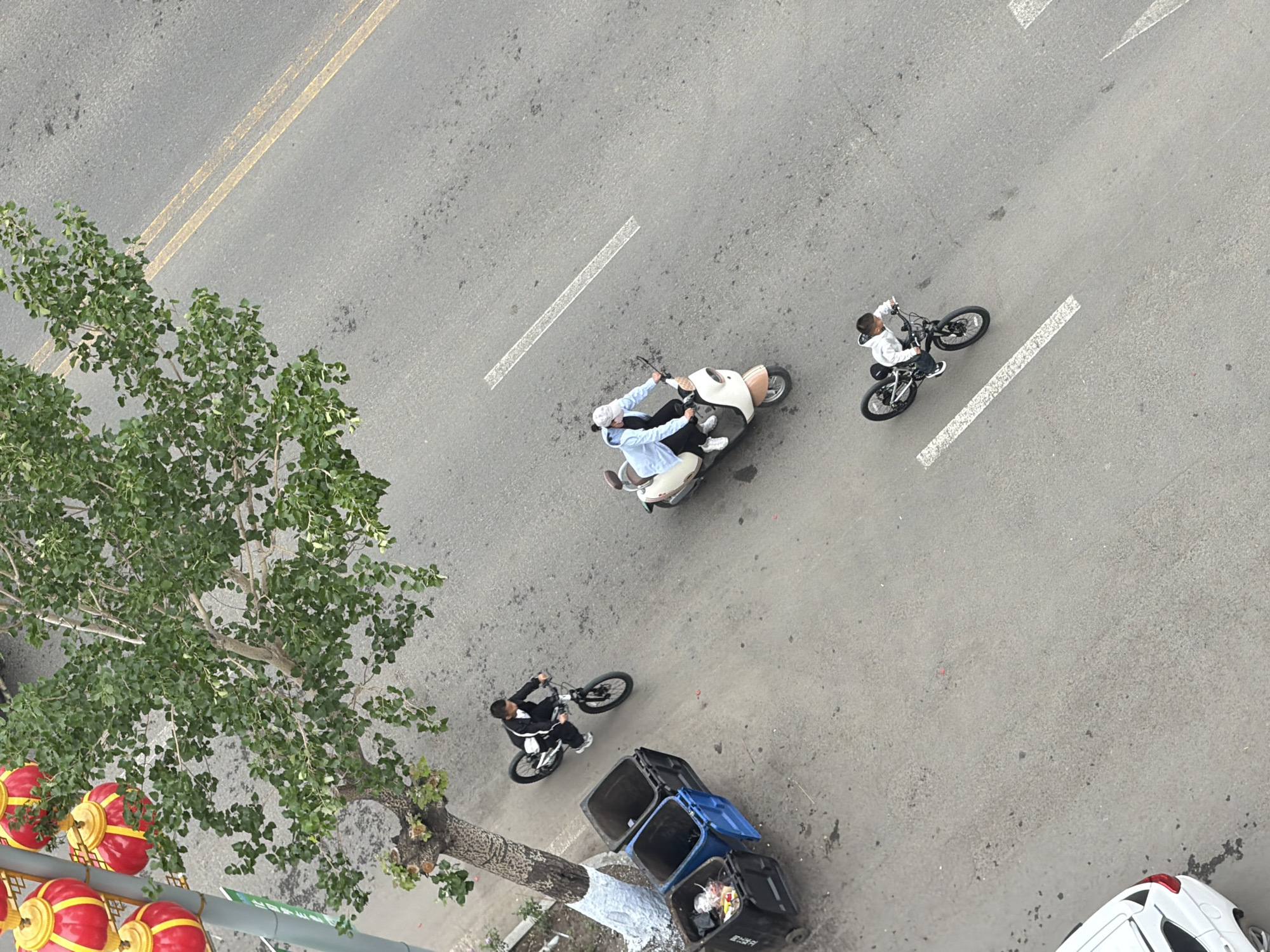 This high-angle overhead shot captures a lively street scene in Tongjiang, China, during daylight hours under clear weather. The grey asphalt road features distinct markings, including double yellow lines, a dashed white line, a solid white line, and white arrow or chevron patterns.
In the mid-ground, two individuals are riding a white scooter or moped. The front rider, dressed in a light blue shirt and a white head covering, steers the vehicle, while a passenger sits directly behind them. To their right, a person in a white hoodie and dark pants is seen riding a bicycle. Further down the street, near the left edge, another individual in a black tracksuit is also riding a bicycle.
A large tree with abundant green leaves dominates the left side of the frame, its branches extending over the street. At the base of the tree, which has a section of its trunk painted white, a series of vibrant red and yellow traditional Chinese lanterns are prominently displayed, hanging from an unseen support structure, adding a strong cultural element to the setting.
On the roadside, near the second cyclist, two large trash bins are visible. One is blue with an open lid, and the other is black, also partially open, showing some refuse inside. Both bins have white Chinese characters inscribed on their sides, likely indicating their respective waste categories. The rear section of a white car is partially visible in the bottom right corner of the image. The scene portrays a routine moment of urban transit and daily life.