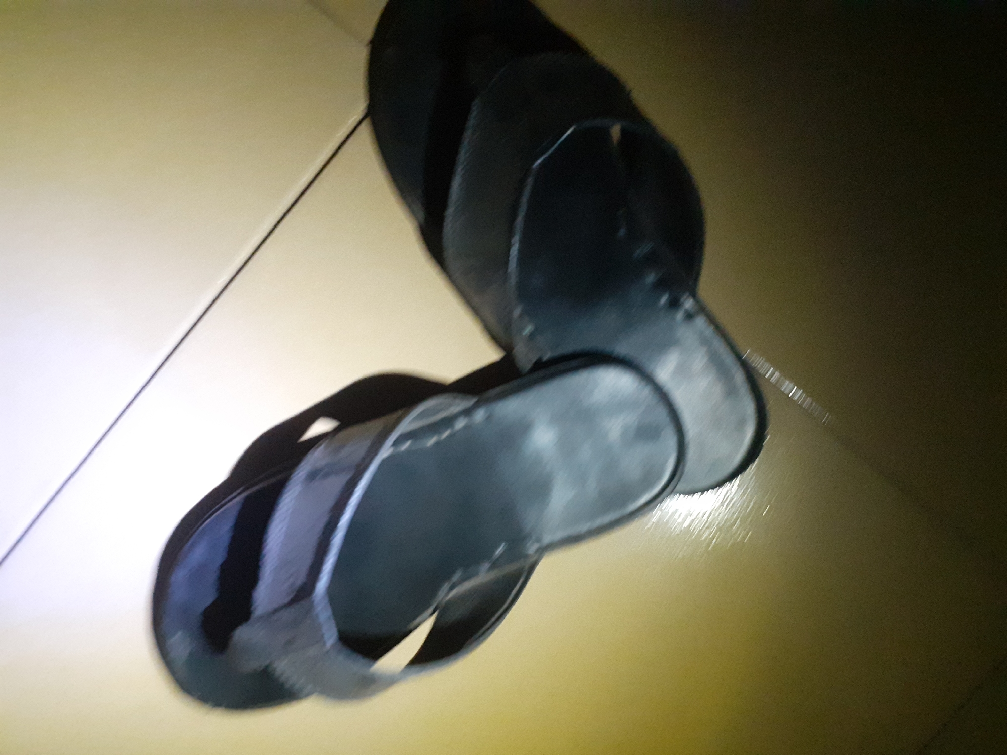 The image shows a pair of black leather flip-flops lying on a light-colored tiled floor. The tiles are arranged in a grid pattern, with a thin dark grout line visible between them. The flip-flops are positioned at an angle, with one slightly overlapping the other. The lighting in the image is uneven, with a bright spotlight creating a harsh glare on the floor and casting shadows of the flip-flops. The background is a plain, light-colored surface, possibly a wall or another section of the floor, also reflecting the light. There are no people visible in the image, and no other objects apart from the flip-flops and the floor. The scene appears to be indoors, possibly in a domestic setting. The location is Jalingo, Nigeria.