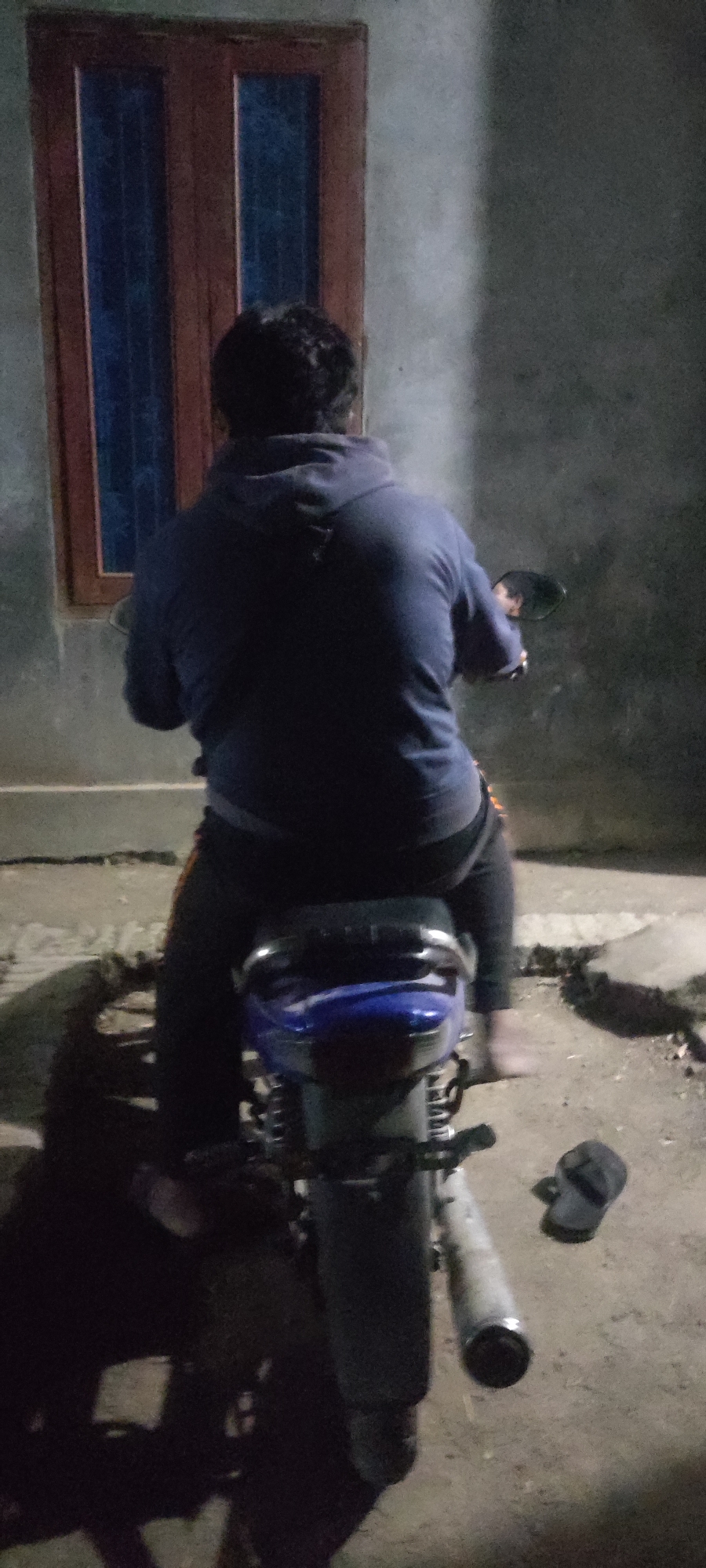 A full shot of a person from behind sitting on a motorcycle at night, with a window visible in the background. The person is wearing a dark blue hooded sweatshirt and dark pants. The motorcycle is blue and black. A pair of flip-flops are on the ground to the right of the motorcycle. The scene is outdoors, with a concrete ground. The lighting suggests it is nighttime or a dimly lit area. The location is Karangwangun, Indonesia.