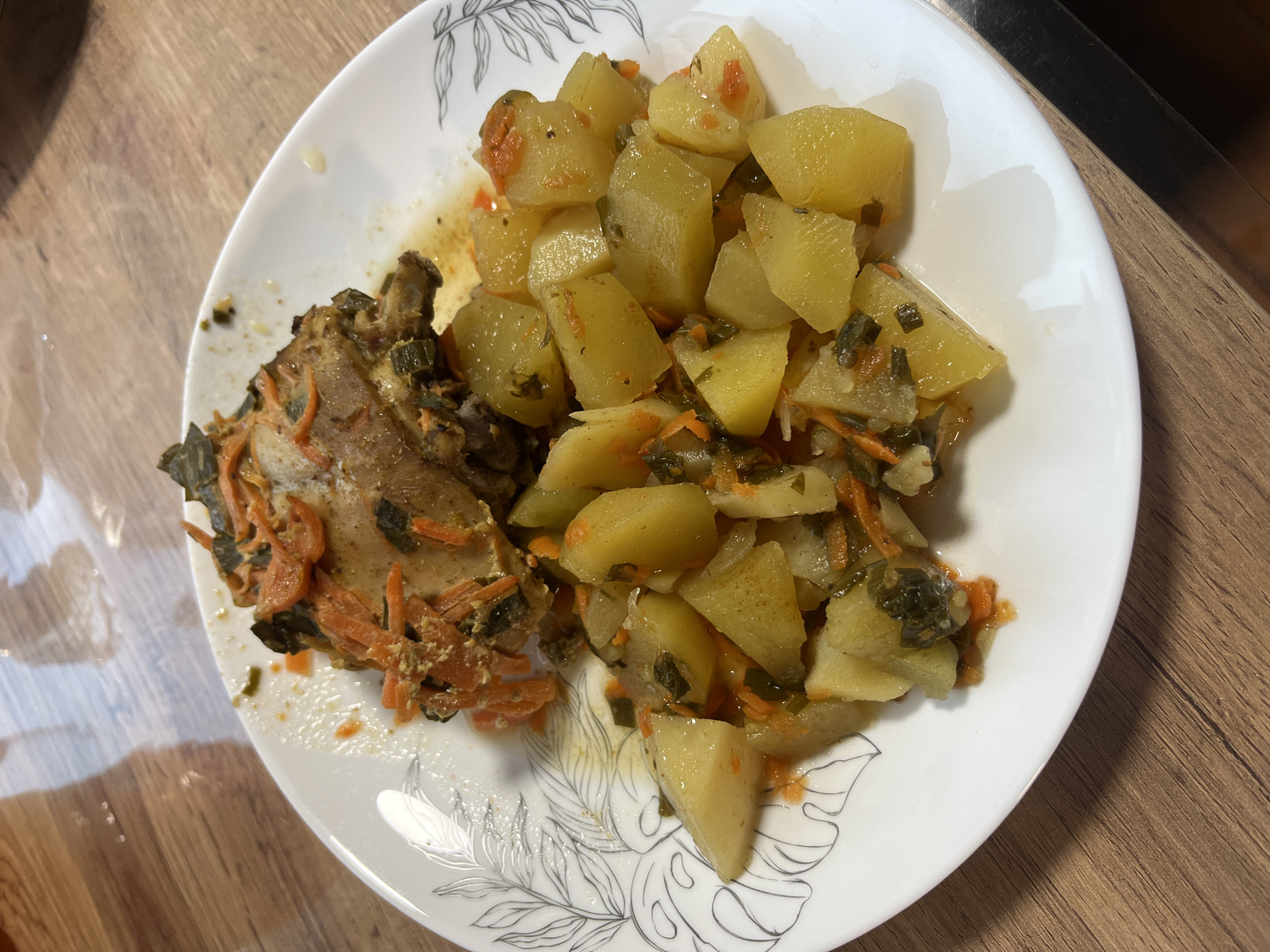 A close-up shot showcases a white plate with a simple black floral design on its rim, filled with a hearty meal. The dish appears to be a stew or casserole, with a prominent piece of meat, likely chicken, on the left side of the plate. The meat is covered in a savory sauce and intermingled with shredded carrots and what look like green herbs or leafy vegetables. To the right of the meat, a generous portion of cubed potatoes is piled high. These potatoes are also mixed with shredded carrots and green vegetables, suggesting they have been cooked together with the meat. The overall impression is of a warm, comforting, home-cooked meal. The lighting is natural, suggesting daylight, and the food is presented on a wooden table, hinting at a casual dining setting. There are no people or text visible in the image. The setting cues are minimal, but the food preparation style suggests a home-style dish.