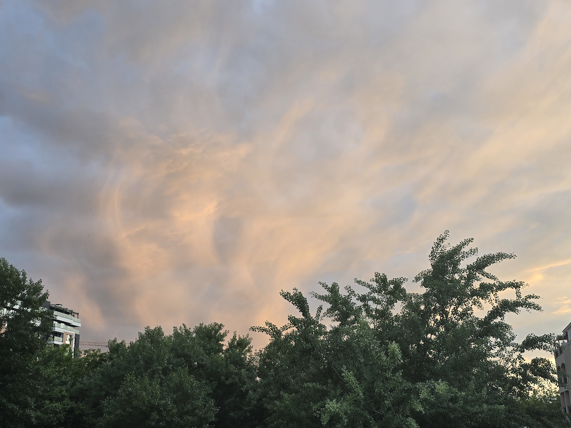 The image shows a sky with a mix of gray and peach-colored clouds, likely at sunset or sunrise. The lower portion of the frame is occupied by green trees, and a building is visible on the left side. The building has multiple stories with balconies, and a construction crane is partially visible behind it. Another building is seen on the right side of the image. The image seems to be taken in an urban environment, likely a city or town.