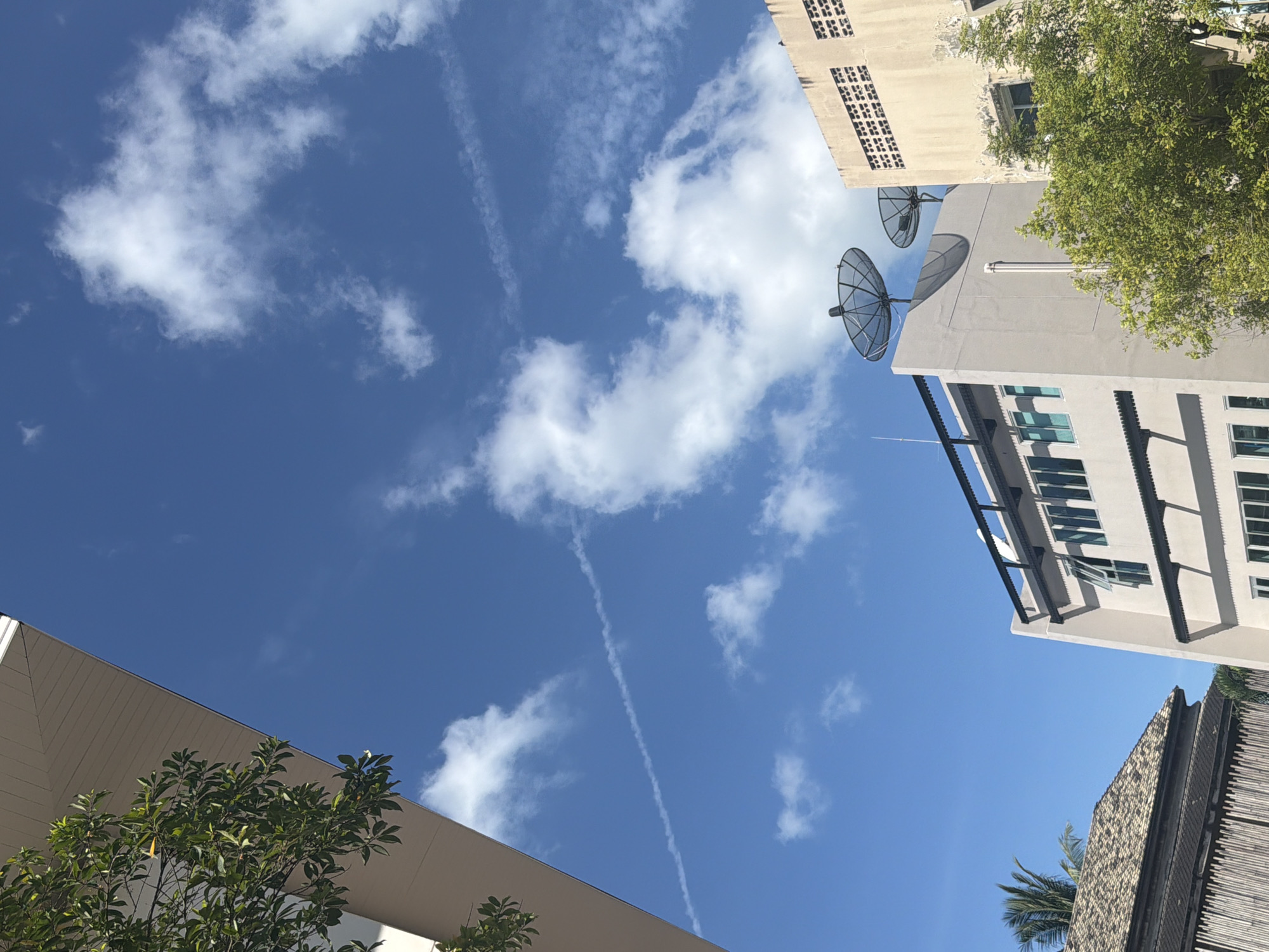 This is a photograph taken from a low angle looking up at a bright blue sky with scattered white clouds. In the sky, there is a faint contrail. In the foreground, the upper portion of a modern building is visible on the left, with green foliage from a tree extending into the frame. To the right, several buildings are visible. One is a light gray building with a black railing along the roofline, and two satellite dishes mounted on top. Adjacent to it is a beige building with a textured facade. Below the gray building, windows with green frames are visible. Further to the left and lower in the frame, a building with a dark, patterned roof and wooden siding is partially visible. The scene suggests an urban or semi-urban environment, possibly in Baan Chaweng, Thailand, given the architectural styles and vegetation. There are no people visible in the image.