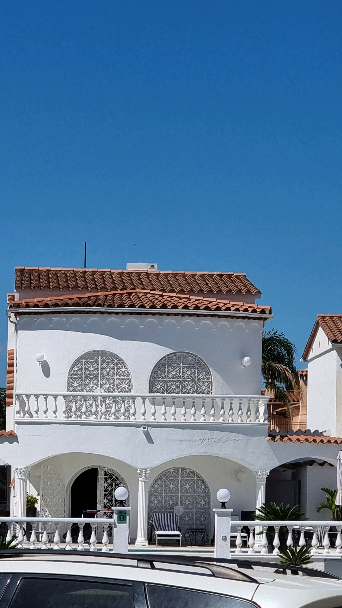 A sunlit, white multi-story house with a red tile roof is visible under a clear blue sky. The house features ornate white balconies and arched windows with decorative metal grilles. In the foreground, the top of a white car is visible. The house appears to be located in a Mediterranean climate, possibly in Castelló d'Empúries, Spain, given the architectural style and the clear, bright sky. There are no people visible in the image. The scene is static and focused on the exterior of the building.