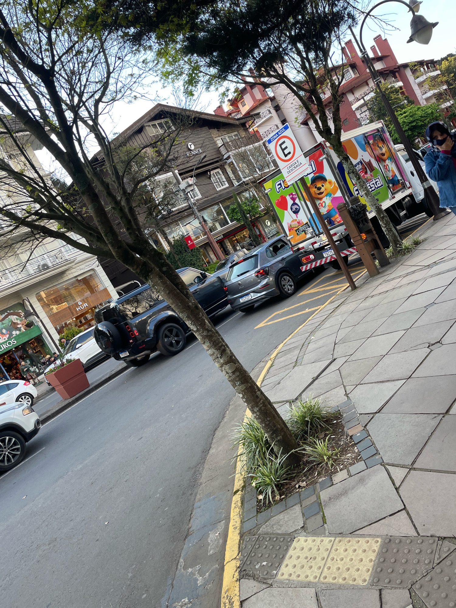 The image depicts a street scene in Gramado, Brazil, likely on Avenida Borges de Medeiros, a popular thoroughfare. The architecture of the buildings suggests a European influence, characteristic of Gramado's style. A dark-colored SUV is parked on the left side of the street, and a grey Volkswagen Polo is behind it. Further down the street, a white truck with colorful cartoon characters on its side is parked, possibly a delivery vehicle. A woman in a denim jacket is standing on the sidewalk to the right, looking towards the camera. Trees line the sidewalk, and streetlights are visible. The sidewalk is paved with hexagonal tiles and features tactile paving for visually impaired individuals. The overall impression is a bustling urban environment, common in a tourist town like Gramado.