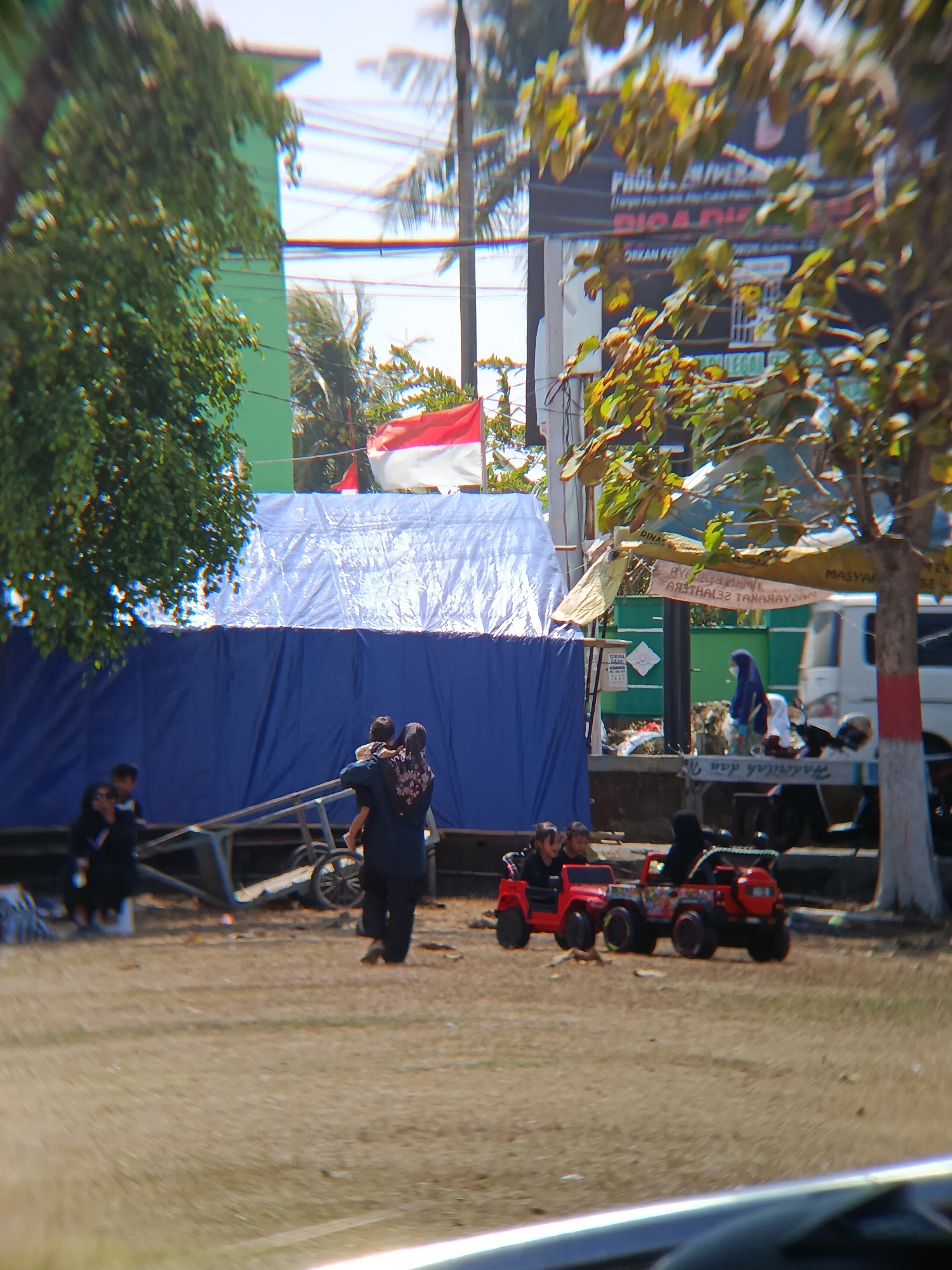 The image depicts an outdoor scene in Desa Kebon Manis, Kecamatan Cilacap Utara, Indonesia. In the foreground, two children are seated in a red toy Jeep, with another child and an adult figure in the background. To the left, several individuals, some in dark clothing, are gathered near a cart. Behind them, a large blue tarp is visible, partially covered by a white tarp. A red and white flag is displayed above the tarps. Further in the background, there are trees, a building painted green, and a billboard with text. A white van and motorcycles are also present in the scene. The ground appears to be unpaved, with scattered debris.