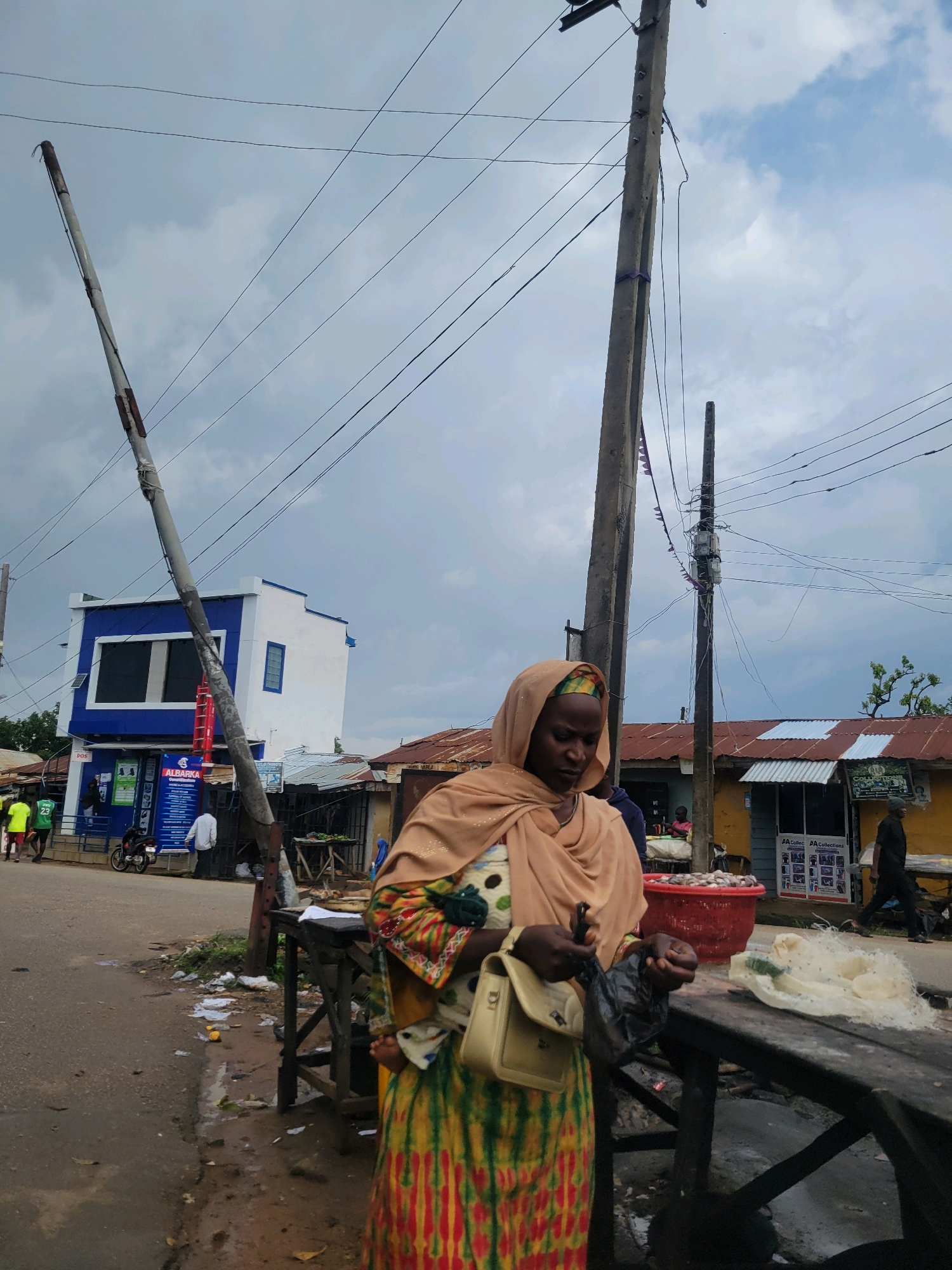 The image depicts a street scene in Jos, Nigeria. In the foreground, a woman wearing a tan headscarf and a colorful dress is holding a black plastic bag and examining its contents. She has a light-colored purse slung over her shoulder. To her right, a wooden table displays some goods, including what appears to be netting and a red basket filled with small items. In the background, a blue and white building with large windows is visible, along with other structures with corrugated metal roofs. Electrical poles and wires crisscross the sky, which is partly cloudy. Several people are walking or standing in the background. The ground appears to be a paved road with some debris and puddles.