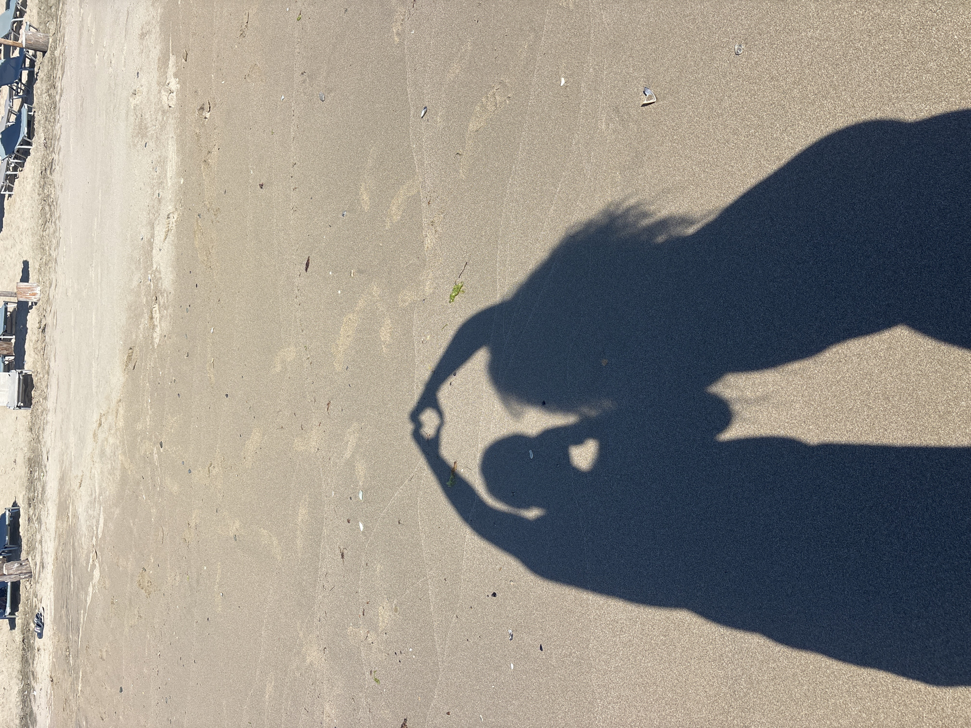 The image shows a shadow of two people forming a heart shape with their hands on a sandy beach. In the background, there are beach chairs and umbrellas. The scene is set in Rezovo, Bulgaria, where the Black Sea meets the mountains. The sand is light brown and textured, with some small shells and debris scattered across it. The shadows are dark and distinct, indicating a sunny day. The overall impression is one of a peaceful and romantic moment captured on a beach.