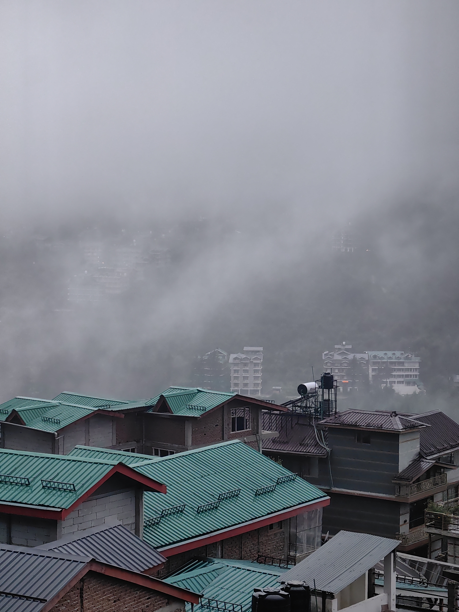 A high-angle, medium shot captures a dense cluster of residential buildings in Vashisht, India, enveloped in thick, atmospheric fog. The foreground is dominated by the colorful, angled rooftops of several houses, predominantly in a vibrant teal or emerald green, outlined with red trim. These structures appear to be constructed with a mix of materials, including brick and light-colored concrete blocks.
Further into the background, the fog obscures the mid-ground and distance, softening the details of larger buildings. These appear to be multi-story hotels or apartment complexes, with their architectural features suggested rather than clearly defined. Some buildings in the mid-ground show hints of balconies and windows, and one prominent structure in the middle features a large water tank and a solar panel system. The distant landscape, visible through breaks in the fog, shows a hilly terrain with more scattered buildings, suggesting the town is built on a slope. The overall impression is one of a serene, slightly mystical, and densely populated urban environment partially hidden by mist. There are no people visible in the frame.