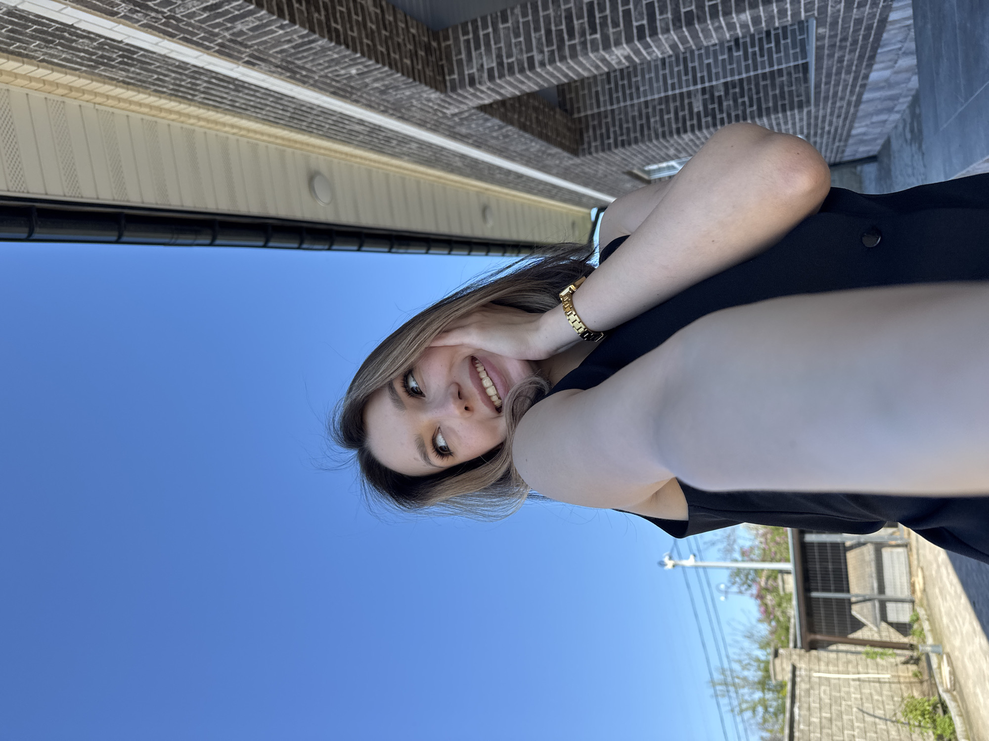 A low-angle view of a fair-skinned woman wearing a black top and a gold watch. She is smiling and resting her head on her hand. Behind her, there is a clear blue sky and a building with brickwork and siding. Further back, there is a partial view of a building with a metal roof and a tree, and a street.