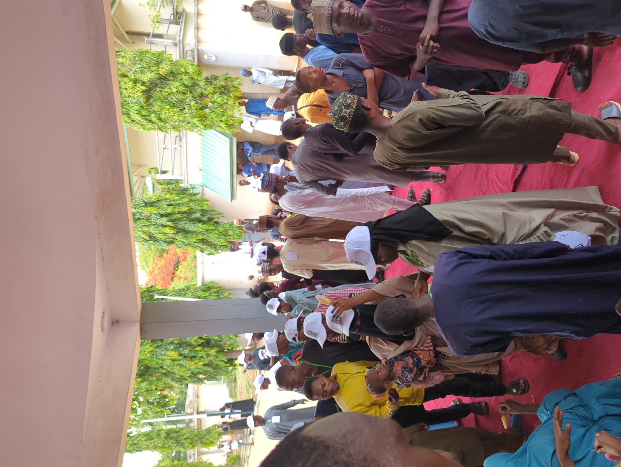 A large group of people are gathered outside under a covered area. They appear to be attending an event or ceremony. The location is Ungwan Liman, Nigeria. The people are dressed in a variety of clothing, including traditional attire and casual wear. Many people are wearing hats, some with logos. Some are seated on a red carpet or mat, and others are standing. The area is partially shaded by a building. There are trees and plants visible in the background, indicating an outdoor setting.