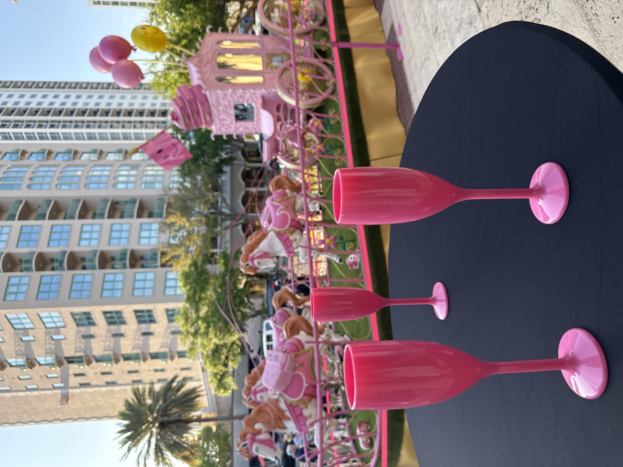 The image captures an outdoor daytime scene in Saint Petersburg, Florida, United States of America, likely on a public promenade or plaza. In the immediate foreground, a round table covered with a dark blue fabric holds three bright pink, opaque plastic champagne flutes, positioned at varying distances from the viewer. The primary subject in the midground is a large, elaborate event float or static display, predominantly pink and gold. This display features multiple decorative pink and white carousel horses with gold accents and saddles, arranged as if in motion. Behind the horses, an ornate pink carriage with gold wheels and illuminated windows is present. A small pink flag with a yellow smiley face is attached to the carriage, and a cluster of pink and yellow balloons (three pink, one yellow) is tethered near its rear. The base of the float is draped in gold fabric and encircled by a decorative pink railing. In the background, tall residential or commercial buildings rise against a clear blue sky. A prominent building on the left is light-colored with numerous balconies featuring green railings and large, light blue-tinted windows with arched tops. A taller, white high-rise building is visible further to the right. Palm trees and other green foliage are scattered throughout the background, and a few indistinct human figures are visible behind the float.