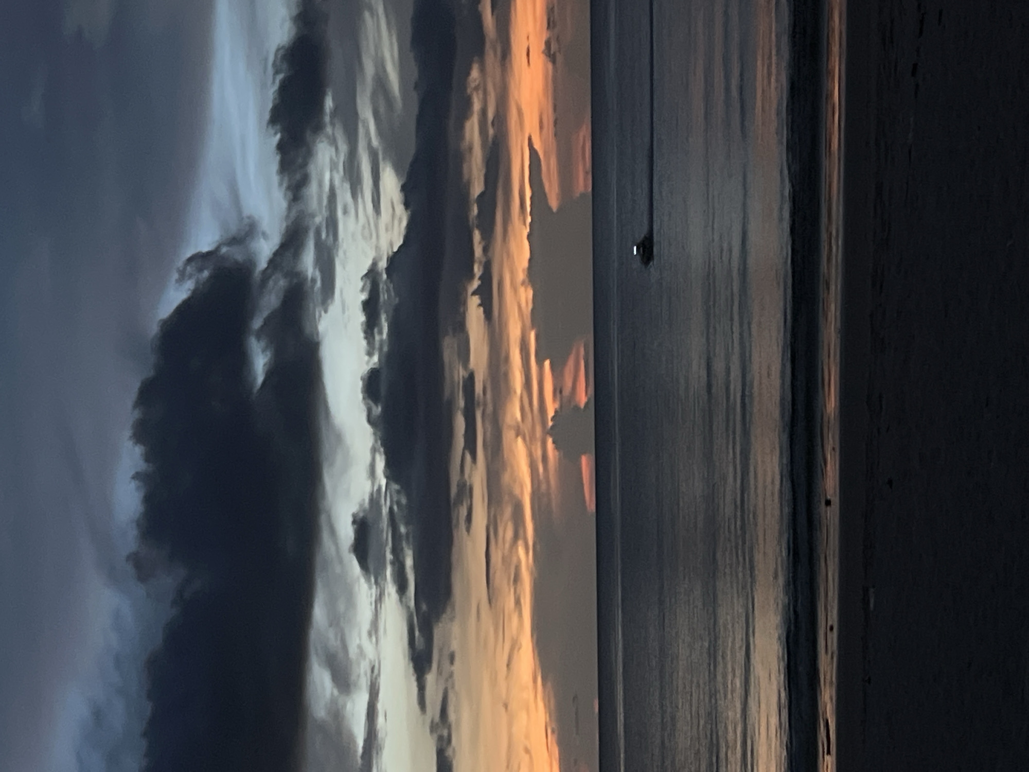 The image shows a seascape at dusk or dawn. The left side of the frame displays dark, stormy clouds, while the right side transitions to orange-tinted clouds reflecting on the water's surface. A long, thin structure pierces the horizon, possibly a pier or pole. The foreground appears to be a dark beach. The location is a beach in an unknown city.