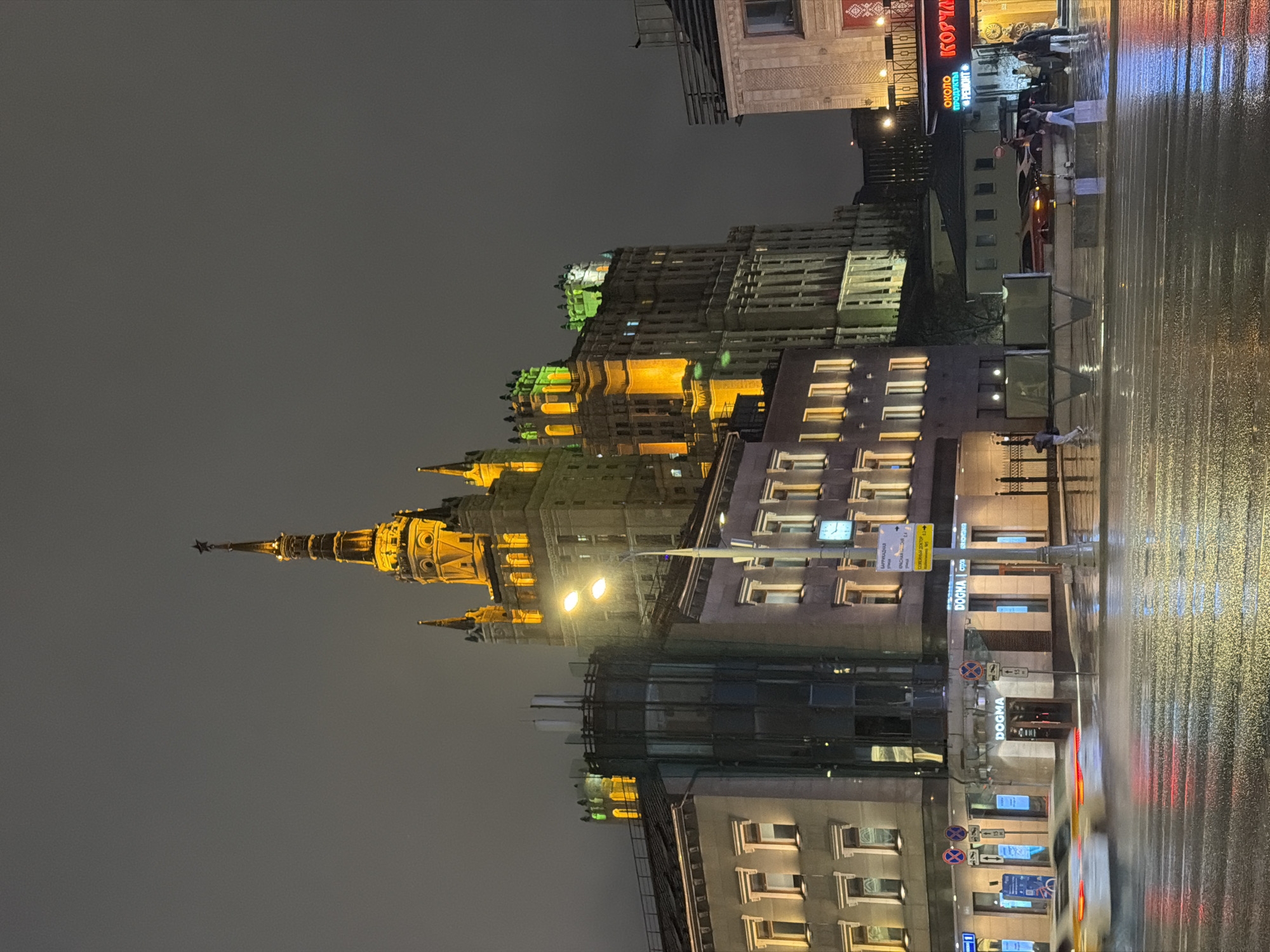 At night, the illuminated Kotelnicheskaya Embankment Building in Moscow, Russia, is seen in a cityscape. The view is captured from street level, with the wet pavement reflecting the city lights. The Kotelnicheskaya Embankment Building stands prominently with its illuminated spire and architectural details. Other buildings line the street, some with lit windows and neon signs. Streetlights and other urban lighting elements contribute to the overall illumination of the scene. Pedestrians can be seen walking, and traffic lights are visible, indicating the presence of vehicles.