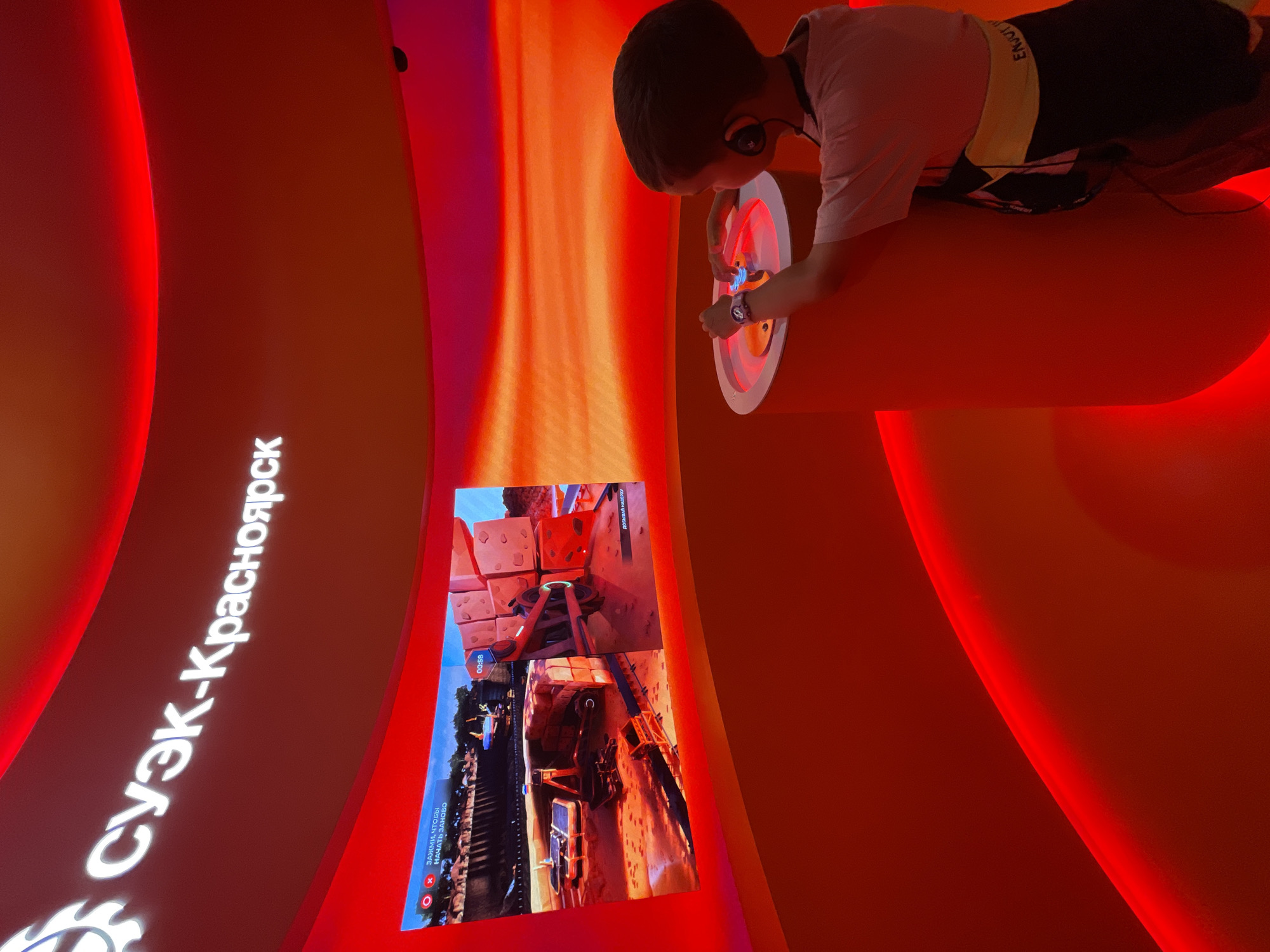 The image depicts an interactive exhibit in Krasnoyarsk, Russia, identified by the Cyrillic text "СУЭК-Красноярск" displayed prominently on a curved, illuminated red wall. The exhibit features a large screen displaying a video game interface with a timer at "00:58" and on-screen text. A young boy wearing headphones and a light-colored t-shirt with a black and yellow trim is actively engaged with a control interface on a circular, illuminated pedestal. He is holding a controller and appears to be playing the game. The environment is characterized by a vibrant, predominantly red color scheme with curved walls and ambient lighting.