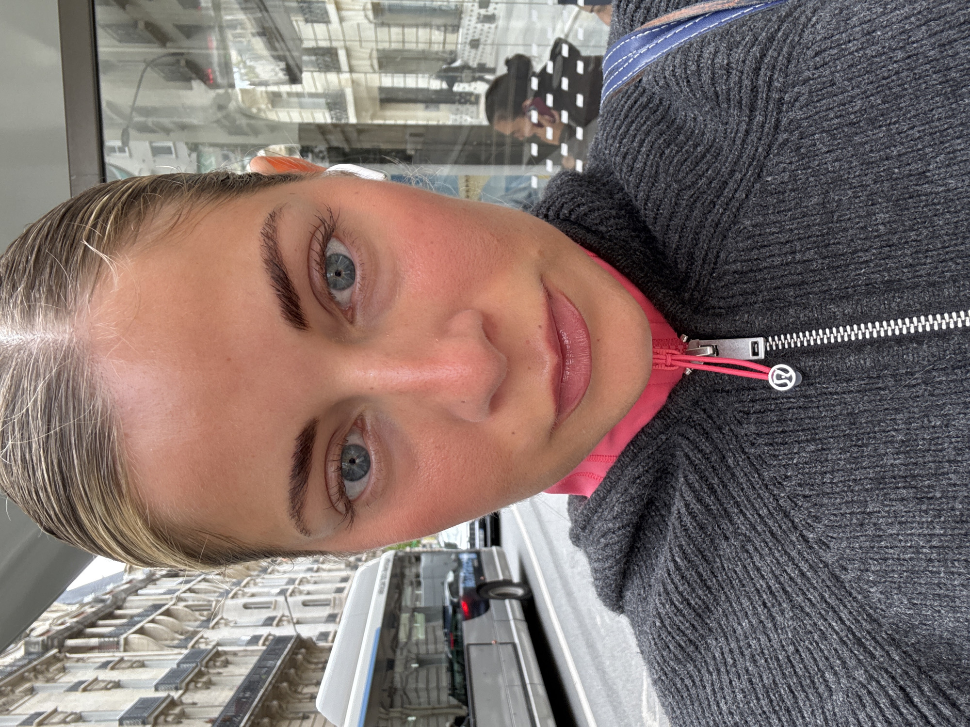 A close-up, slightly angled headshot captures a woman with light blonde hair and blue eyes. She is wearing a grey knit zip-up sweater with a pink inner lining. An earbud is visible in her left ear. The background, visible through a window or reflection, suggests an urban environment with light-colored buildings featuring multiple windows. A bus is partially visible in the street. Based on the architectural style, this scene may be in Paris, France.
