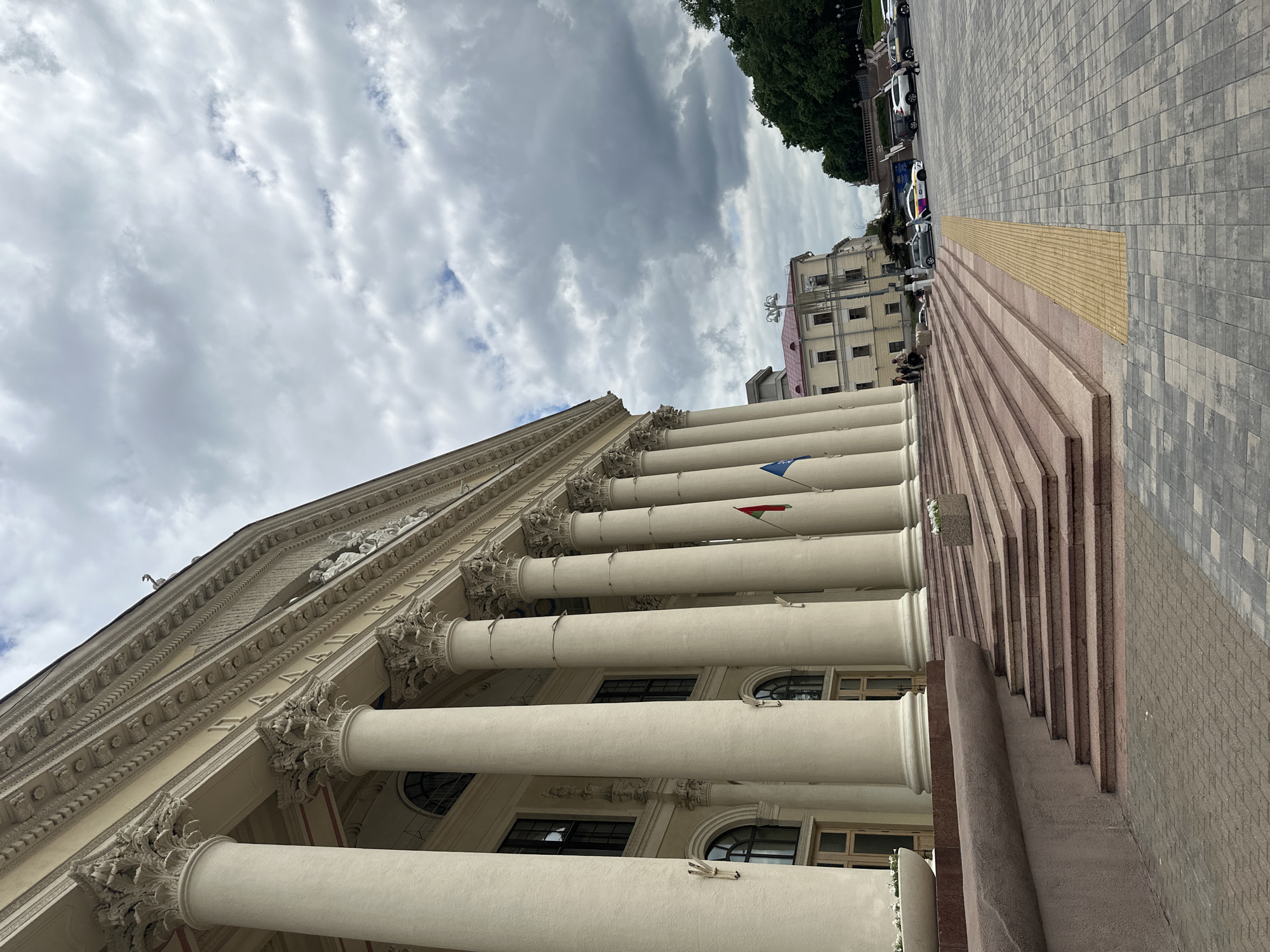 The image shows the Grand Staircase of the Palace of Republic in Minsk, Belarus, under a cloudy sky. The monumental building, constructed in the neoclassical style, features a series of massive Corinthian columns supporting a long entablature. The foreground is dominated by wide, multi-tiered steps made of red granite. To the left, a low, curved concrete wall with planters containing white flowers lines the base of the staircase. The ground in front of the stairs is paved with grey and beige interlocking bricks, with a contrasting yellow tactile paving strip running parallel to the steps.
Several people are visible on the stairs and in the plaza. Some individuals are walking, while others are standing or sitting near the entrance. A few cars are parked in the background, including a white SUV and a yellow taxi. Two flags, one red and white and the other blue, are planted near the entrance of the building. In the background, other buildings with red roofs and trees can be seen. The overall atmosphere is that of a public space on a somewhat overcast day, with the grandeur of the Palace of Republic as the central focus.