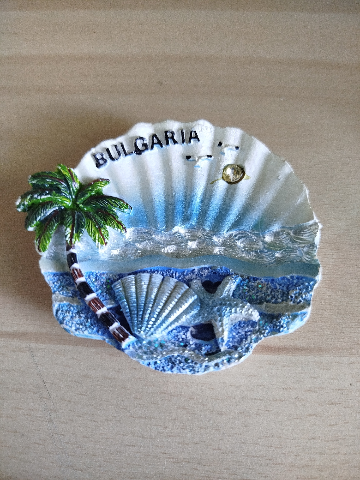 A souvenir magnet, shaped like a seashell, rests on a light brown, wood-like surface. The magnet depicts a coastal scene, featuring a palm tree with green leaves and a brown trunk on the left side. In the center, there is a white seashell and a starfish on a bed of blue, glittery "sand." Above the sand is a layer of "water" with silvery wave effects. The top portion of the magnet resembles the inside of a shell, painted white with blue streaks, and the word "BULGARIA" written in black letters along the top edge. There is also an image of two seagulls and the sun.