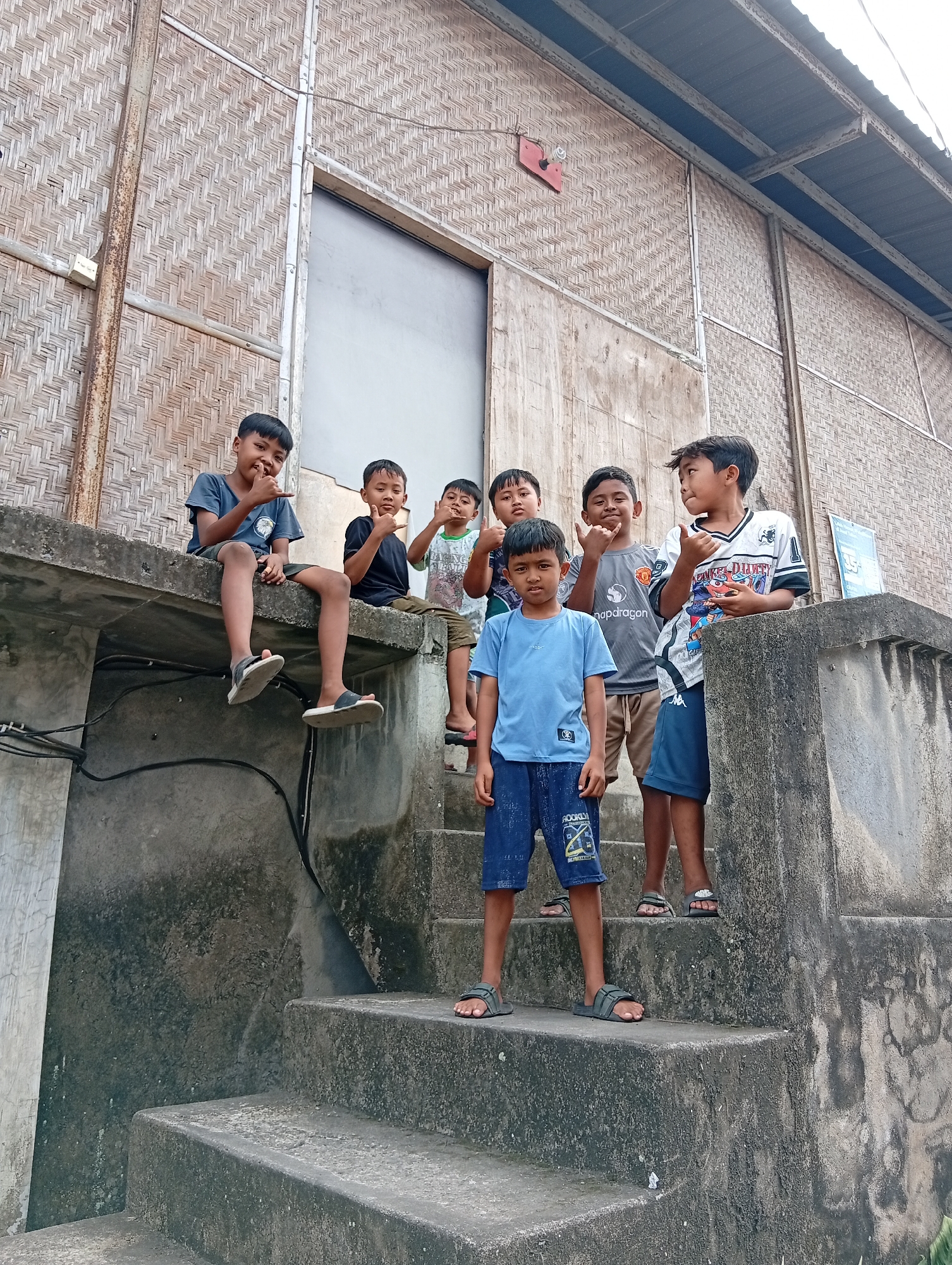 In Ubud District, Indonesia, a group of seven young boys are gathered outside a building with a woven bamboo facade. The boys, appearing to be between the ages of 6 and 12, are positioned on concrete steps and a ledge. One boy sits on the ledge in the foreground, facing left, wearing a dark blue t-shirt and shorts. To his right, another boy sits on the ledge, looking towards the camera, dressed in a black t-shirt and brown shorts. Behind them, on the steps, a boy in a light green and white t-shirt is partially visible. Further to the right and slightly down the steps, a boy in a grey t-shirt with a logo is giving a thumbs-up. Next to him, a boy in a white and black jersey with colorful graphics is also giving a thumbs-up. In the front, standing on the steps, is a boy in a light blue t-shirt and dark blue shorts, looking directly at the camera. The environment is textured with rough concrete and the distinct pattern of the woven bamboo wall. The overall scene captures a candid moment of childhood outdoors in a distinctly Indonesian setting.