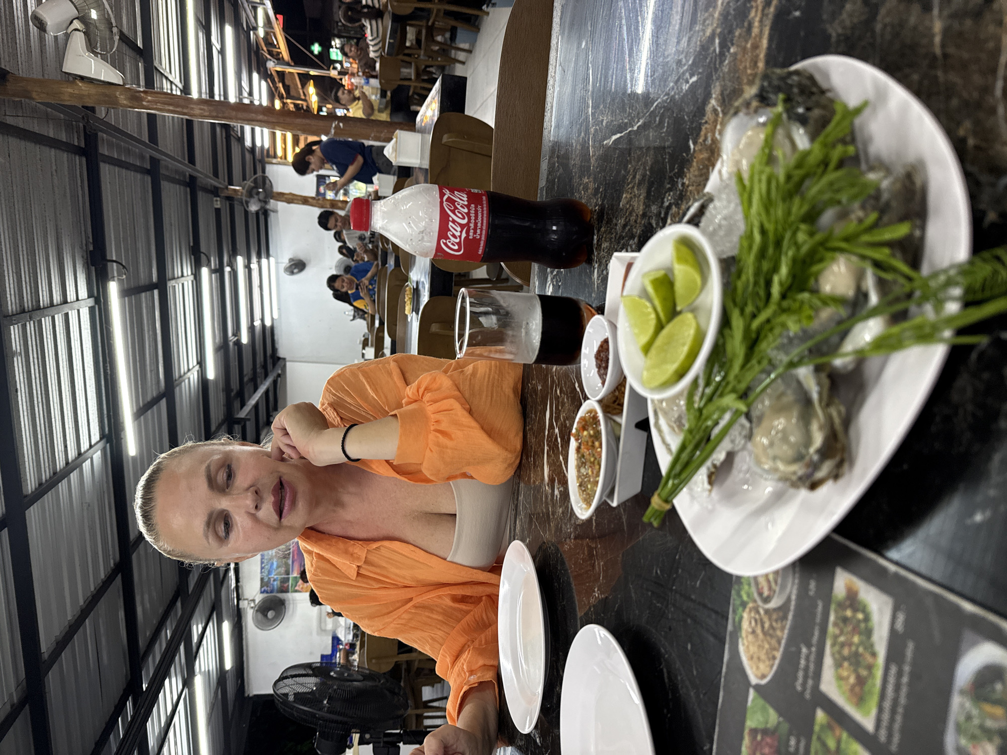 A woman with blonde hair pulled back is seated at a table in an outdoor restaurant on Koh Samui, Thailand, at night. She is wearing an orange, open-necked shirt and a beige top, with her left hand resting on her chin. In front of her, a black marble table displays a plate of oysters with lime wedges and green herbs, two white plates, a glass of dark liquid, a bottle of Coca-Cola, and small bowls of condiments. The background shows other diners and the restaurant's ceiling with metal beams and fluorescent lighting. A large black fan is visible to the left of the woman. The overall scene suggests a casual dining experience.