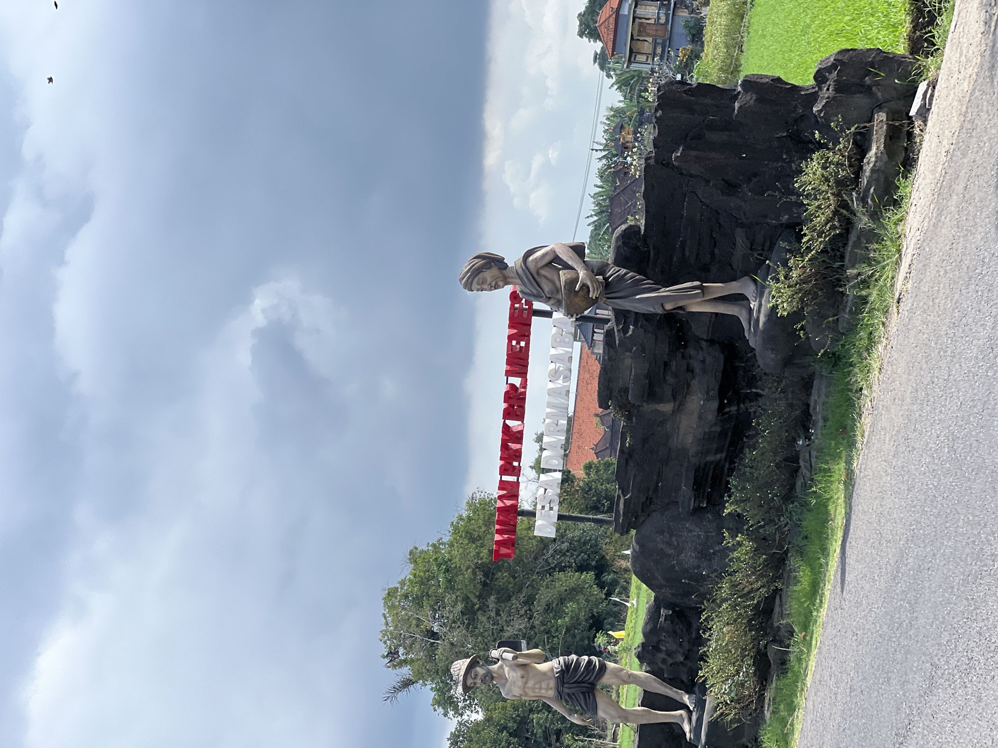 The image captures a scene in Abiansemal, Indonesia, featuring two statues on dark, rocky bases. The statue on the left depicts a bare-chested man wearing a hat and a sarong, holding a farming tool. The statue on the right portrays a woman in traditional attire, carrying a bowl. Behind the statues, a sign displays text in red and white, with "DESA DARMASABA" clearly visible in white letters on the lower line. The background includes lush green rice fields, traditional Indonesian houses with red tiled roofs, and a cloudy sky. A paved road runs along the foreground.