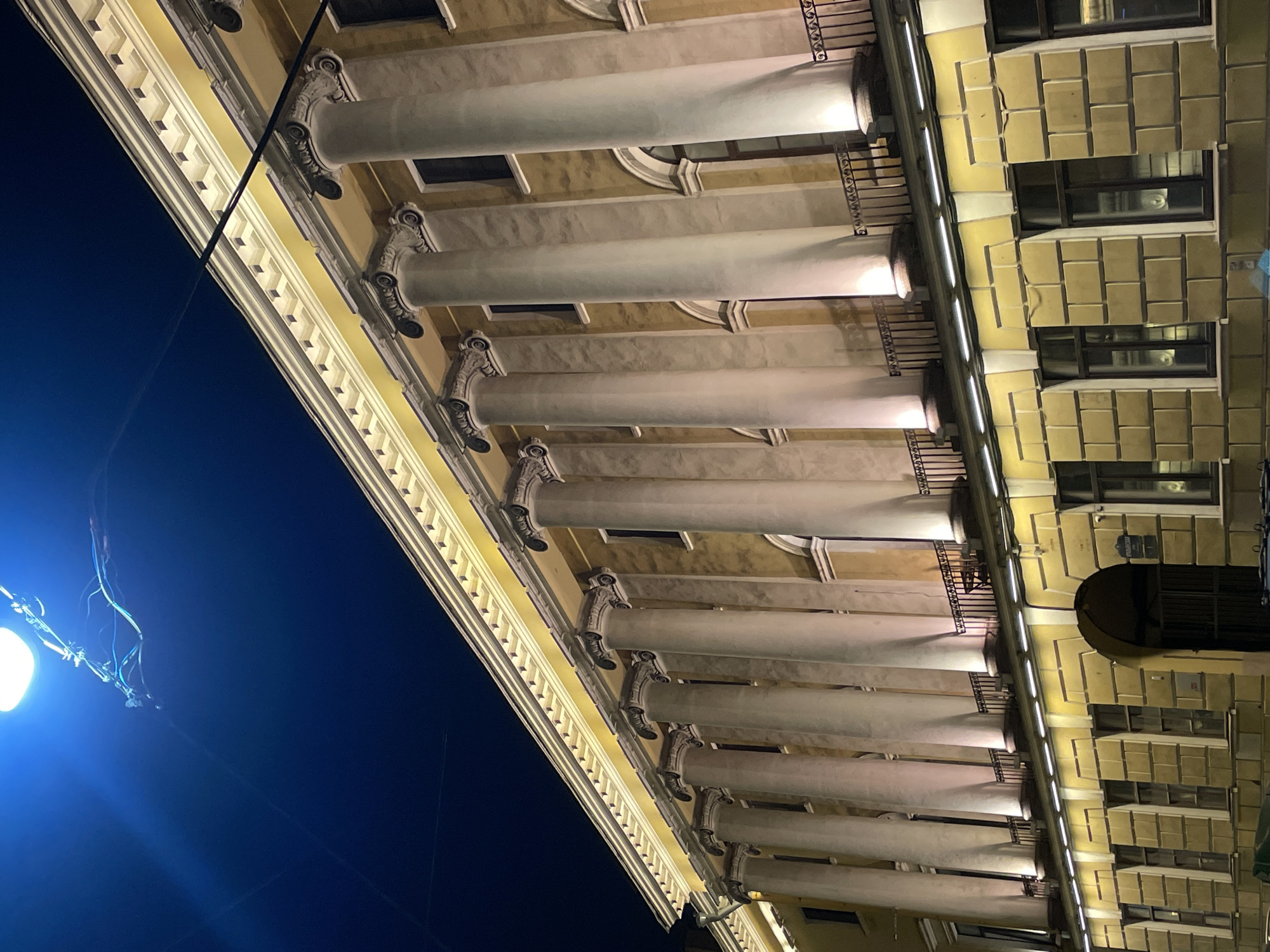 The image shows the facade of a building in Saint Petersburg, Russia, viewed from below at night. The building features a series of columns supporting a balcony and a decorative cornice. The sky is dark blue, and a street lamp illuminates the scene. The building's windows are visible.