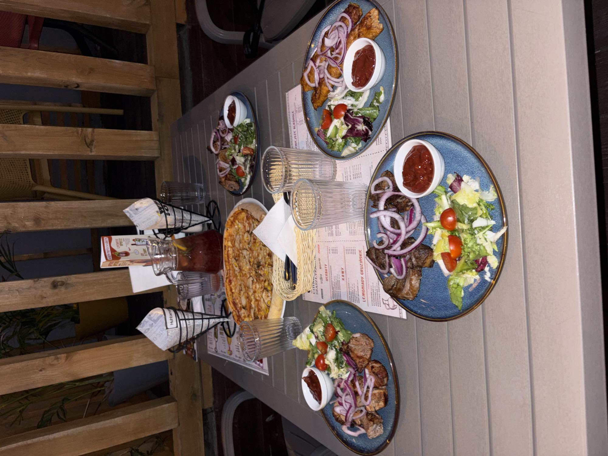 The image shows a table setting at an outdoor restaurant in Lublin, Poland. On the table, there are three plates of food, each containing grilled meat, a side salad with tomatoes and lettuce, and a small bowl of ketchup. There is also a pizza, a pitcher of iced tea, and several glasses. In the background, there are wooden structures, plants, and chairs. The overall scene depicts a meal being served at a restaurant.