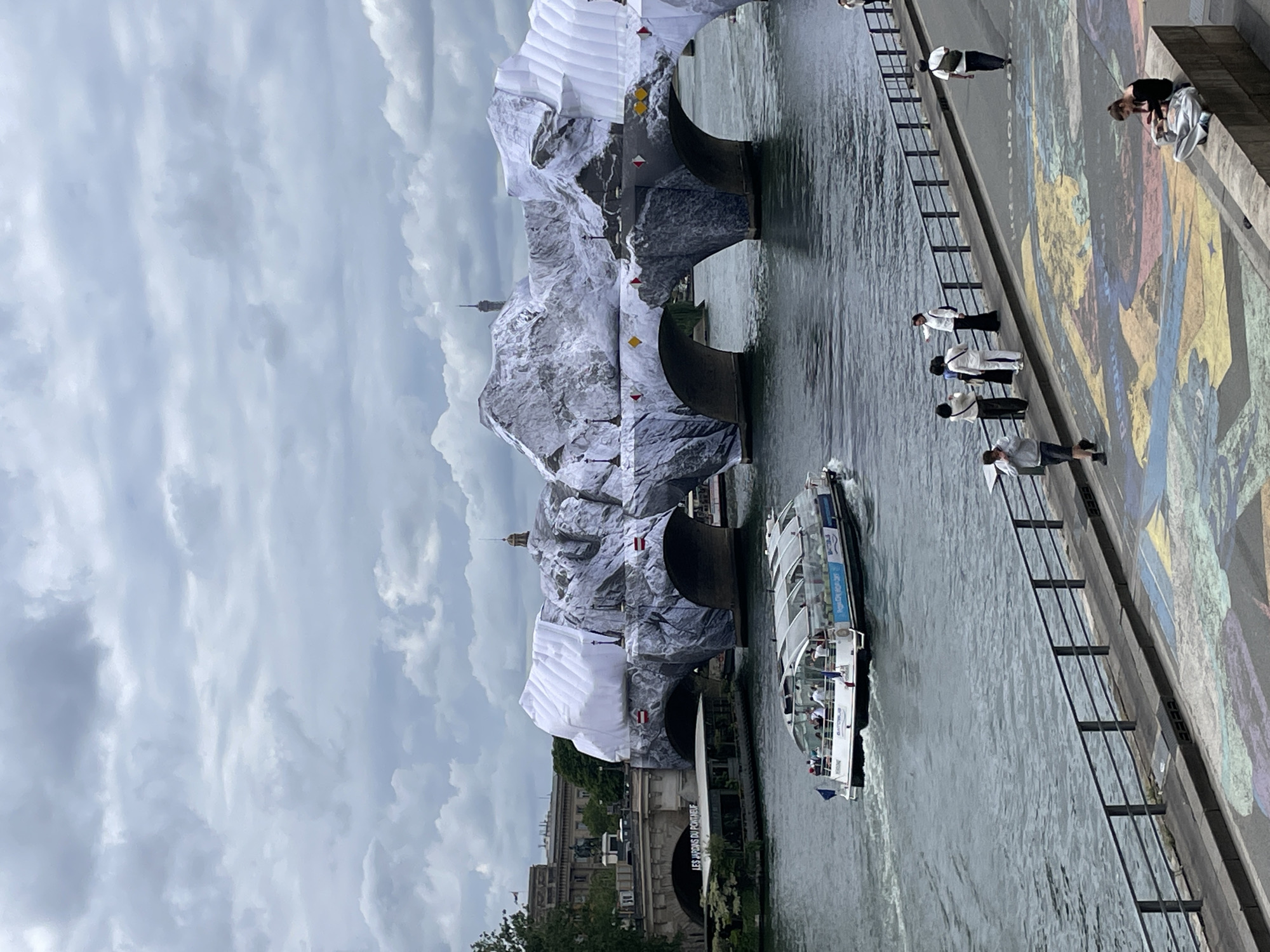 An exterior, eye-level, full shot captures a wide view of the Pont Neuf in Paris, France. The bridge is adorned with large, white inflatable sculptures resembling icebergs, with images of icy terrain printed on them. A tour boat filled with passengers navigates the Seine River in the foreground, moving from left to right. On the riverbank to the right, several people are visible, some standing and looking at the bridge, while one person sits on a ledge, reading. The ground in the foreground features a colorful mural. The sky is overcast with heavy clouds, and the Eiffel Tower is faintly visible in the distance behind the bridge.