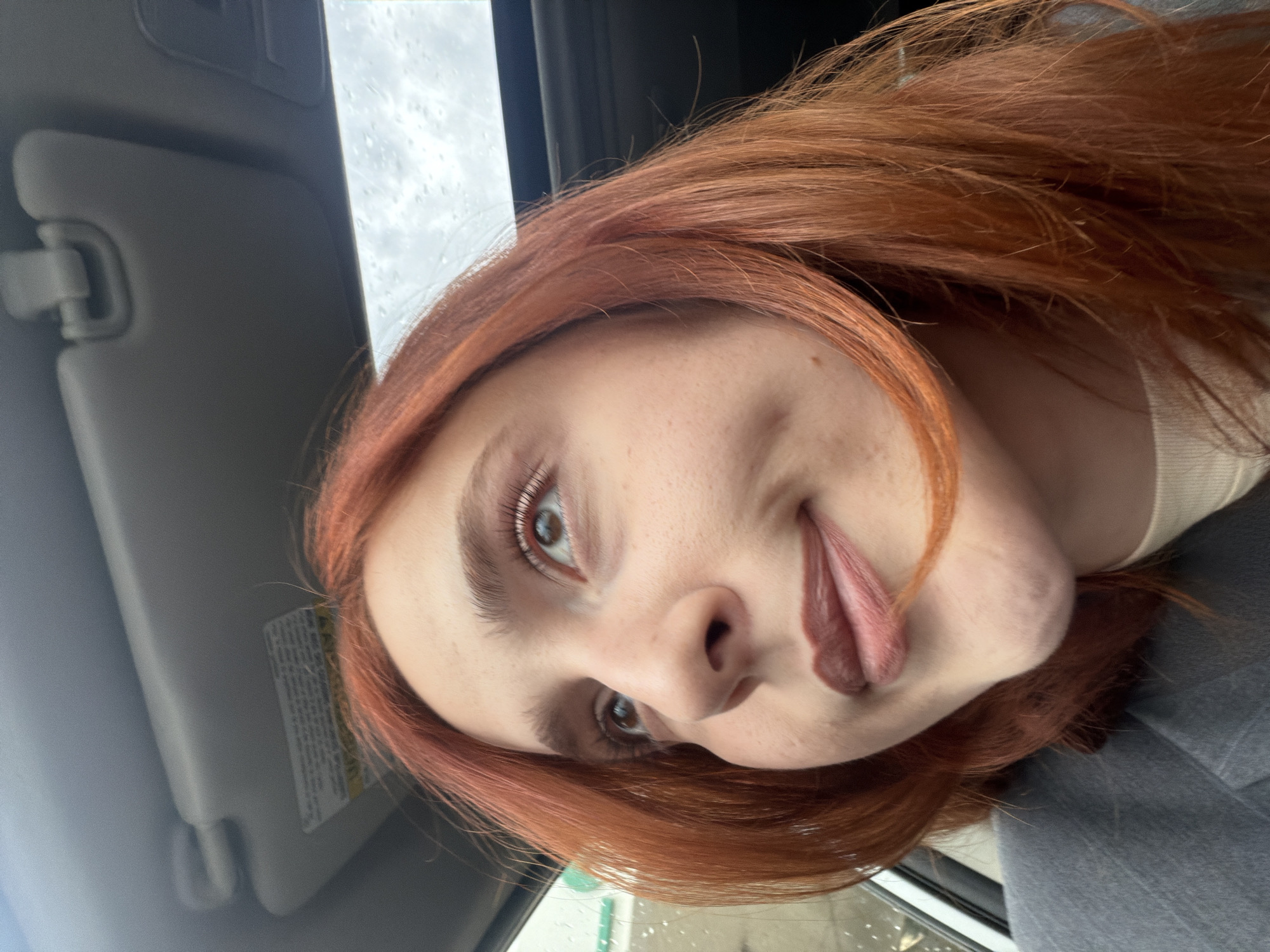 A person with reddish-brown hair is in a vehicle, potentially a car, as indicated by the sun visor. They are sticking out their tongue. Rain appears to be falling on the windows. The person's clothing consists of a light-colored top and a grey jacket or sweater. The location cannot be determined as Tbilisi, Georgia without additional visible indicators in the image.