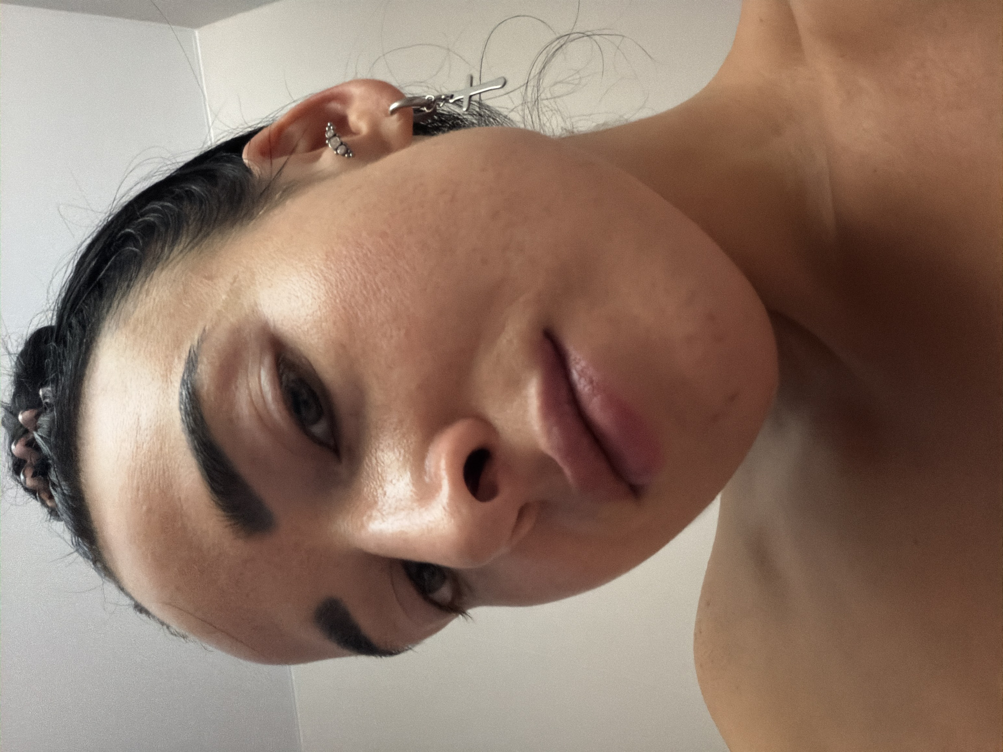 A close-up, rotated image captures the face and upper torso of a young woman from a low angle. Her head is tilted, and she looks directly towards the camera with a neutral expression. Her dark hair is pulled back, with some wisps visible near her left ear.
Notable details include her prominent, dark eyebrows and full, slightly parted lips. On her left ear, there are two distinct piercings: one is a curved bar featuring three small, light-colored beads or stones, positioned on the outer cartilage. The second piercing holds a dangling silver earring in the shape of a cross. Her skin appears natural, showing some texture.
The woman's bare shoulder and collarbone are visible, suggesting she is either topless or wearing a strapless garment. The background is a plain, light-colored wall, out of focus. The lighting is bright and even, indicative of an indoor setting. There is no visible text in the image.