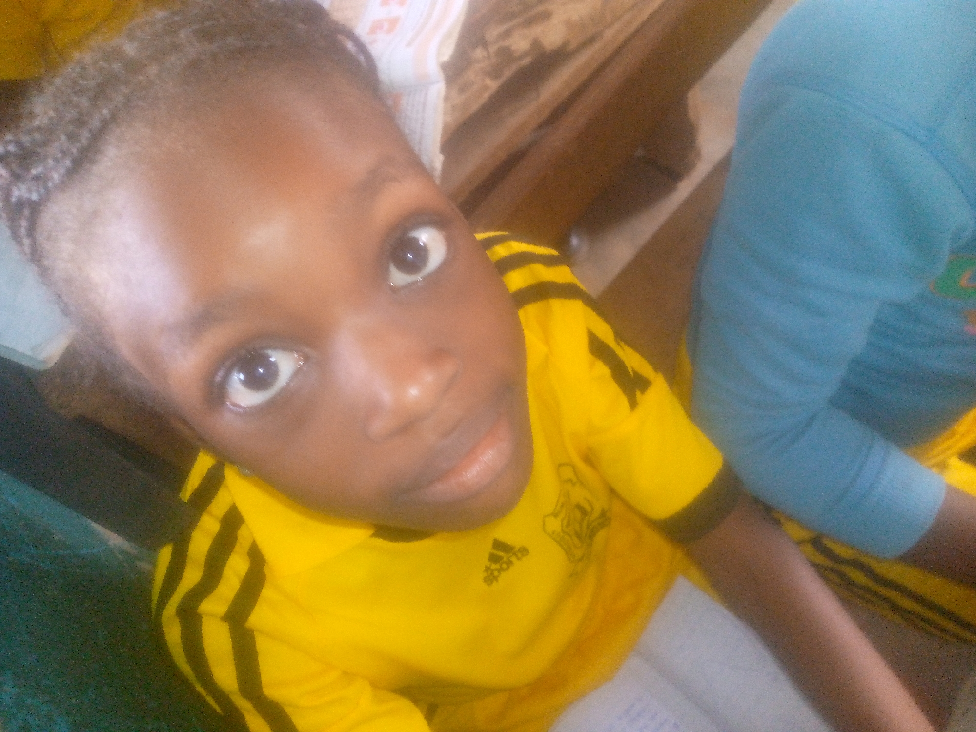 A close-up shot captures a young, brown-skinned person wearing a yellow shirt with black stripes. The Adidas logo is visible on the shirt. Another person wearing a light blue shirt is partially visible in the upper right corner. They are both sitting at a desk. A book or stack of papers are in the background, along with a wooden shelf or desk. The location is unknown.