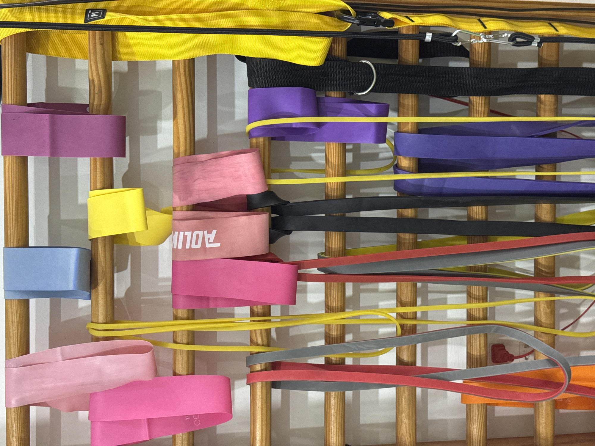 The image depicts a close-up, eye-level shot of a collection of colorful resistance bands and straps hanging on a wooden gym ladder against a white wall. The bands are of various colors including pink, light blue, purple, yellow, red, and gray, and are of different widths and lengths. Some are looped over the wooden rungs of the ladder, while others hang down vertically. A black strap with a metal ring and a yellow strap with a carabiner clip are also visible on the right side of the image. The scene is well-lit, with the focus on the variety and organization of the exercise equipment. The location is identified as Nam Nha Trang Ward, Vietnam.