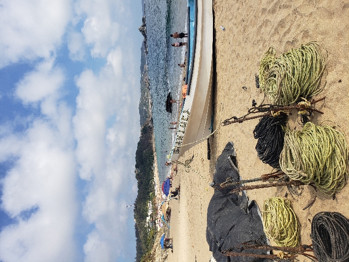 A beach scene with a white boat resting on the sand near the shore. Several bundles of rope, both light and dark colored, are scattered beside the boat. People are visible in the water and on the beach in the distance. The sky is filled with fluffy white clouds. A coastal hillside with trees can be seen along the beach.