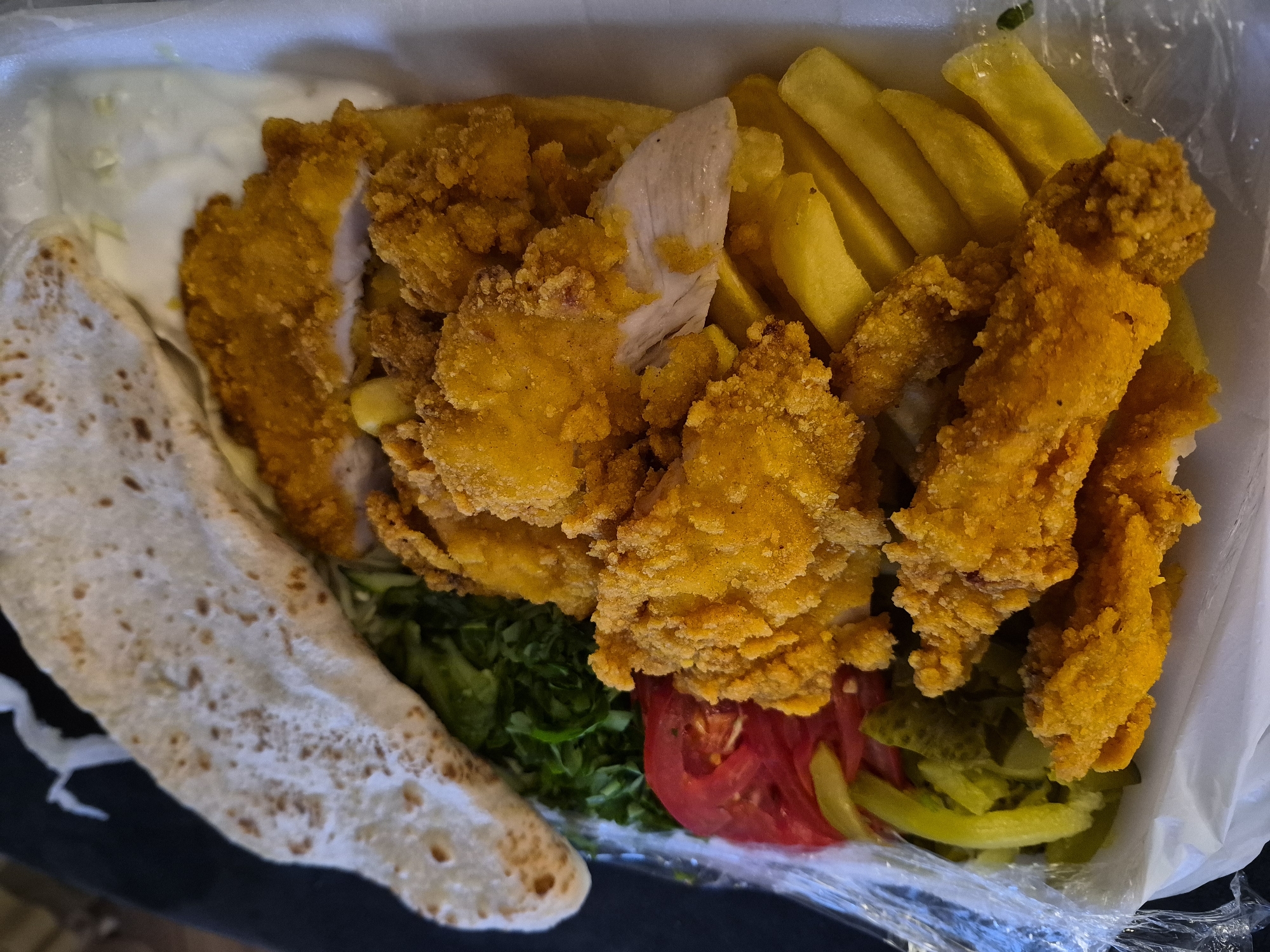 A high-angle, close-up shot captures a takeout meal presented in an open, white, rectangular disposable foam container. The central focus is a generous portion of golden-brown, breaded fried chicken pieces, several of which are broken or sliced, revealing the white cooked meat inside. Interspersed with and beneath the chicken are numerous thick-cut, golden-yellow french fries. To the upper left, a folded, light-colored flatbread, marked with small brown specks, rests on a bed of fresh green leafy vegetables, likely lettuce or parsley. Adjacent to the greens are several slices of vibrant red tomato and an array of green pickled items, including what appear to be sliced cucumbers and peppers. This section of the meal is partially covered by a thin layer of clear plastic wrap. A creamy white sauce is visible in the upper right quadrant of the container, behind the fried chicken. The background is dark and out of focus, providing no specific environmental details. The overall scene depicts a prepared meal for consumption, likely from a fast-food establishment or restaurant. The location is Unknown, Unknown.