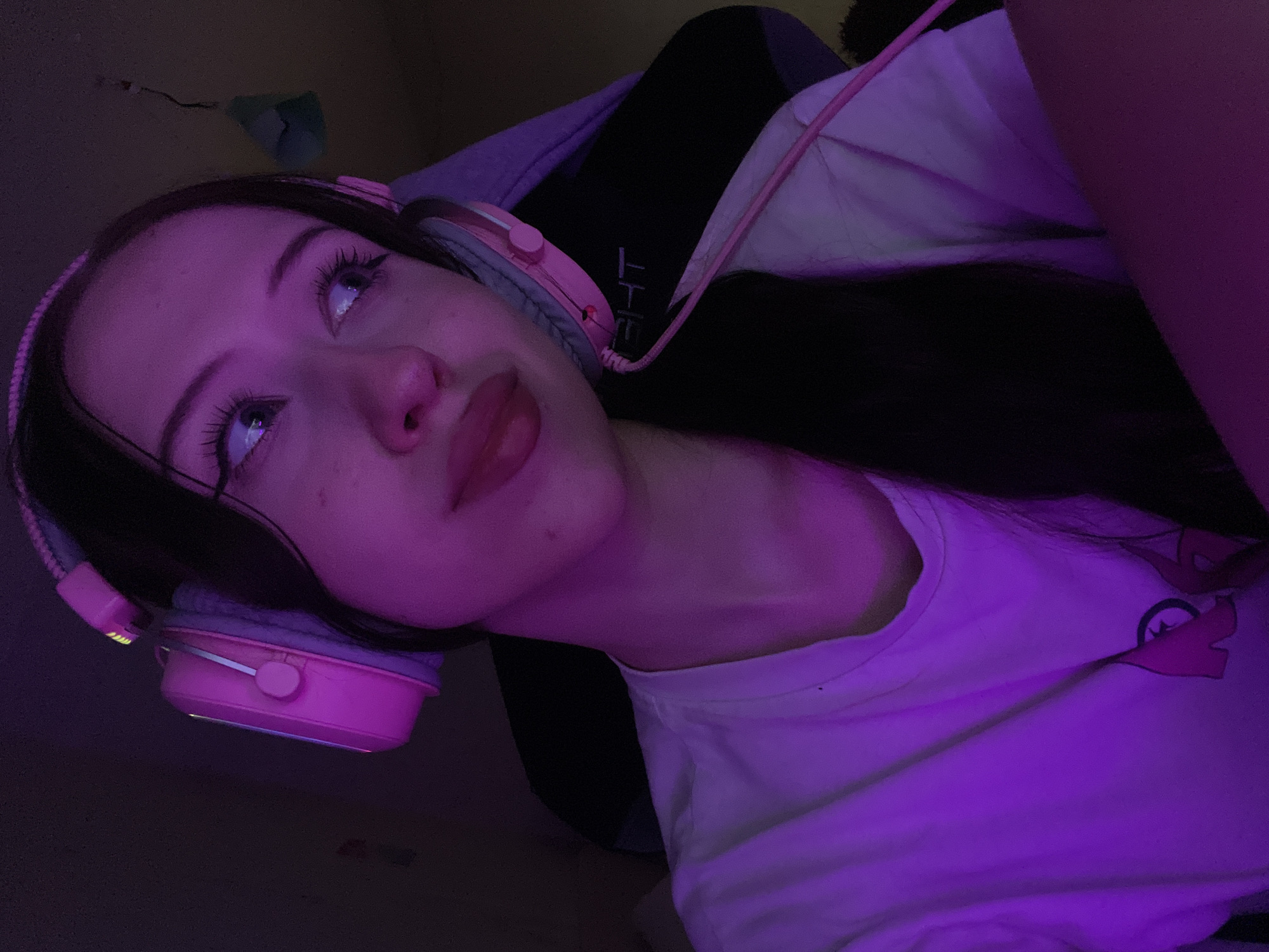 An over-the-shoulder, medium shot captures a young woman with long, dark hair wearing pink headphones and a white t-shirt, illuminated by purple lighting. She is seated in a dark gaming chair, with a portion of the chair's headrest visible behind her. The woman is looking off to her right with a slight smile, her makeup featuring prominent winged eyeliner and full lips. The overall scene is dimly lit, with the purple hue creating a vibrant, atmospheric effect. There is no indication of location in Votkinsk, Russia.
