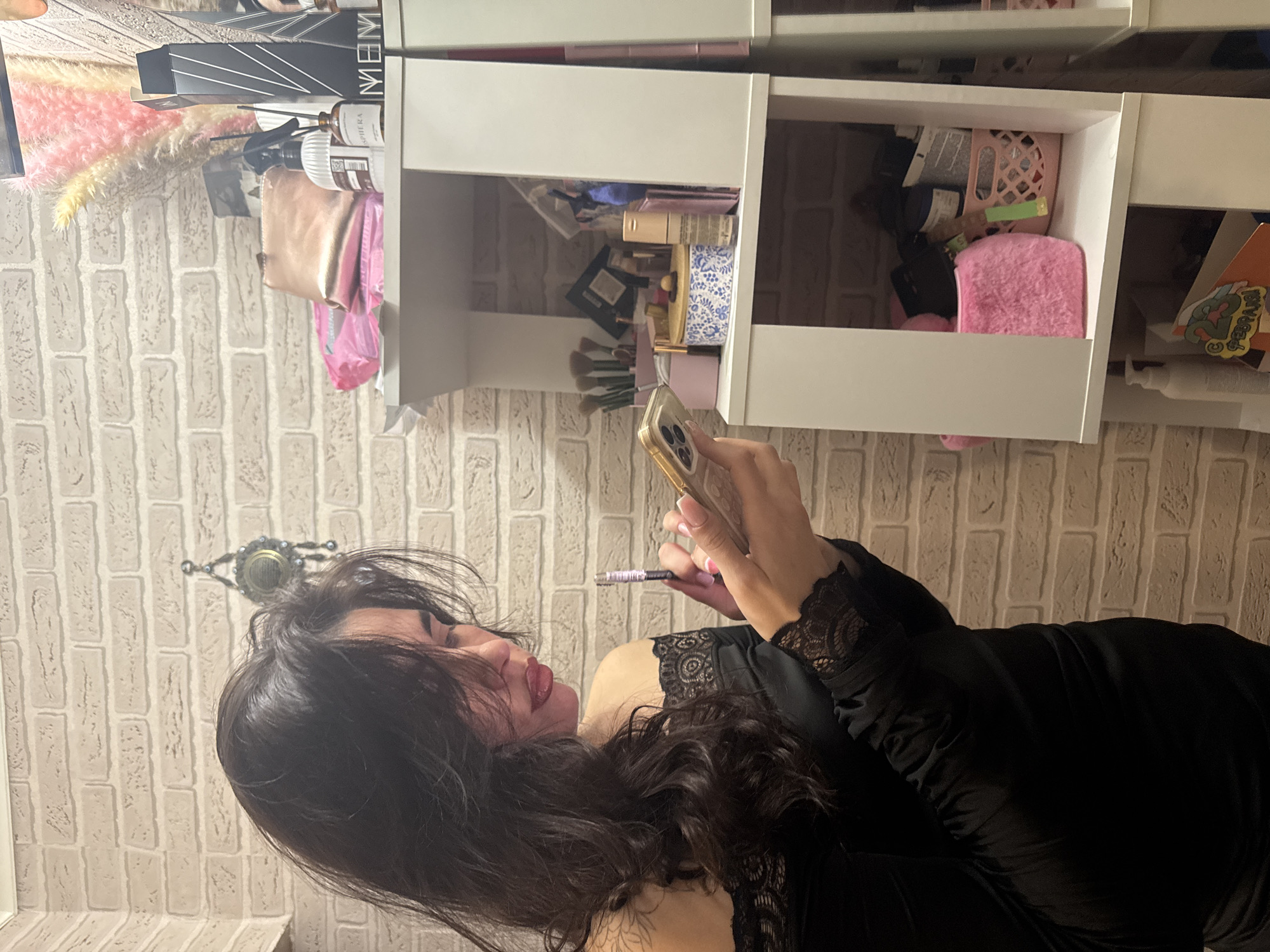 A woman with long dark hair is holding a smartphone in her left hand and a makeup pencil in her right hand. She is wearing a black dress with lace details. She is standing in front of a set of white shelves filled with various items, including makeup, beauty products, and accessories. The background is a brick wall. The location is most likely in a home setting in the city of Chaikovsky, Russia.