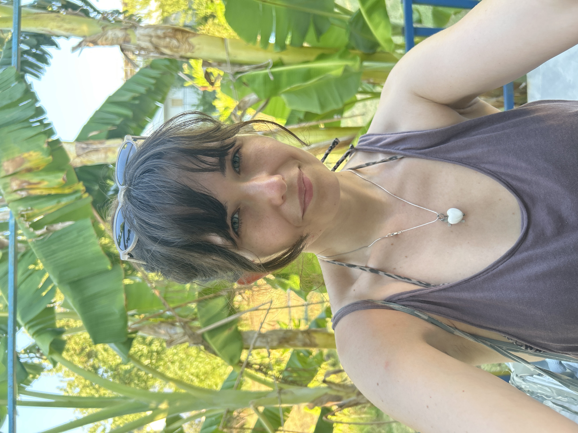 A young Caucasian woman with dark hair and blue-green eyes is taking a selfie outdoors in Ortaca, Turkey. She is wearing a dusty purple tank top and a necklace with a white heart pendant. Sunglasses are perched on top of her head. She is smiling slightly and looking at the camera. Behind her, banana trees with large green leaves are abundant. The background also shows hints of trees and a building. The lighting suggests it is daytime, with sunlight filtering through the foliage. A blue railing is visible on the right side of the frame.