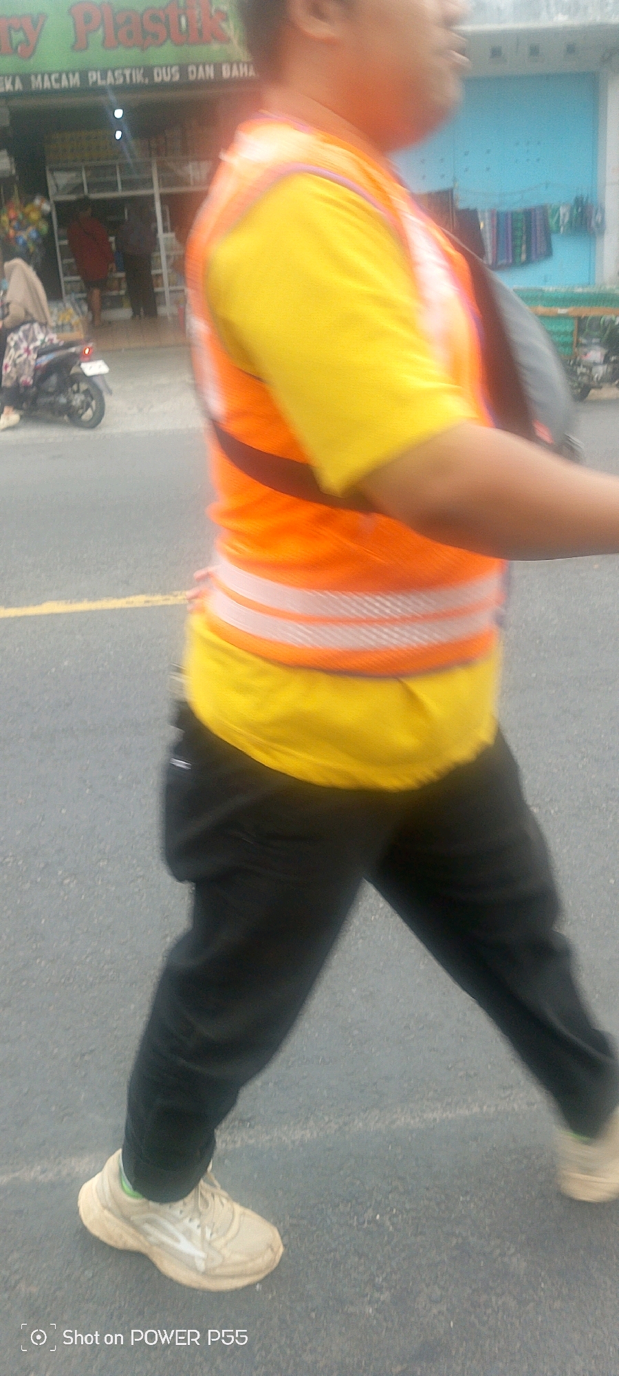 A person is walking on a road. They are wearing a yellow t-shirt, an orange vest with reflective stripes, and black pants. They have on light-colored sneakers. In the background, there is a storefront with a sign that reads "EKC MACAM PLASTIK, DUS DAN BAHA". There are also motorcycles and buildings visible in the background.