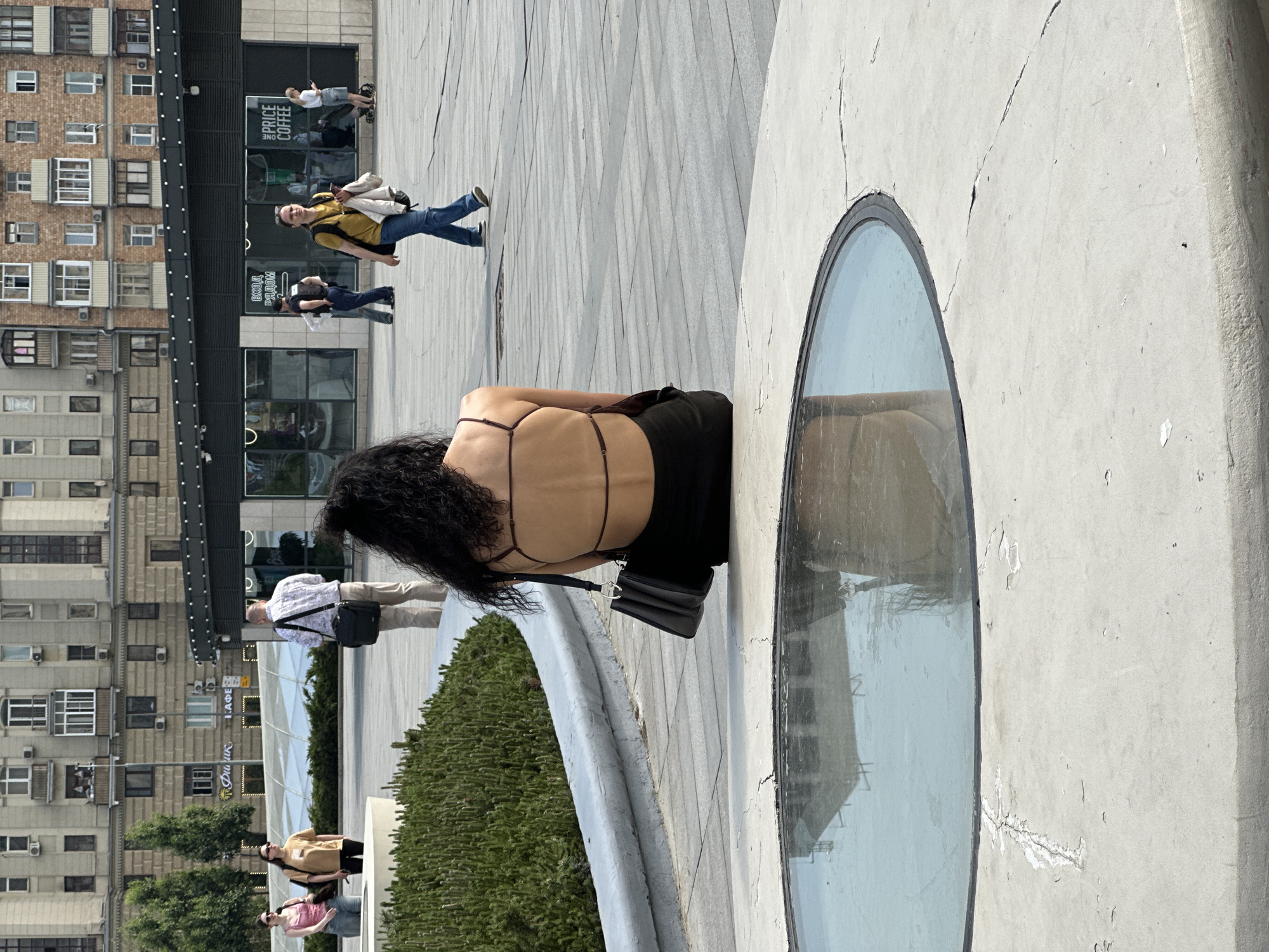 The image shows an outdoor urban scene, likely in a public plaza. A woman with dark curly hair, wearing a black top and pants, stands near a circular structure with a glass-covered opening, possibly a skylight. A black purse hangs from her shoulder. The structure is surrounded by greenery. In the background, several other people are walking. Behind them is a building with numerous windows, likely an apartment complex, along with storefronts at the ground level. The location appears to be Moscow, Russia. The plaza has light-colored paving stones with a patterned design. The weather seems sunny and bright.