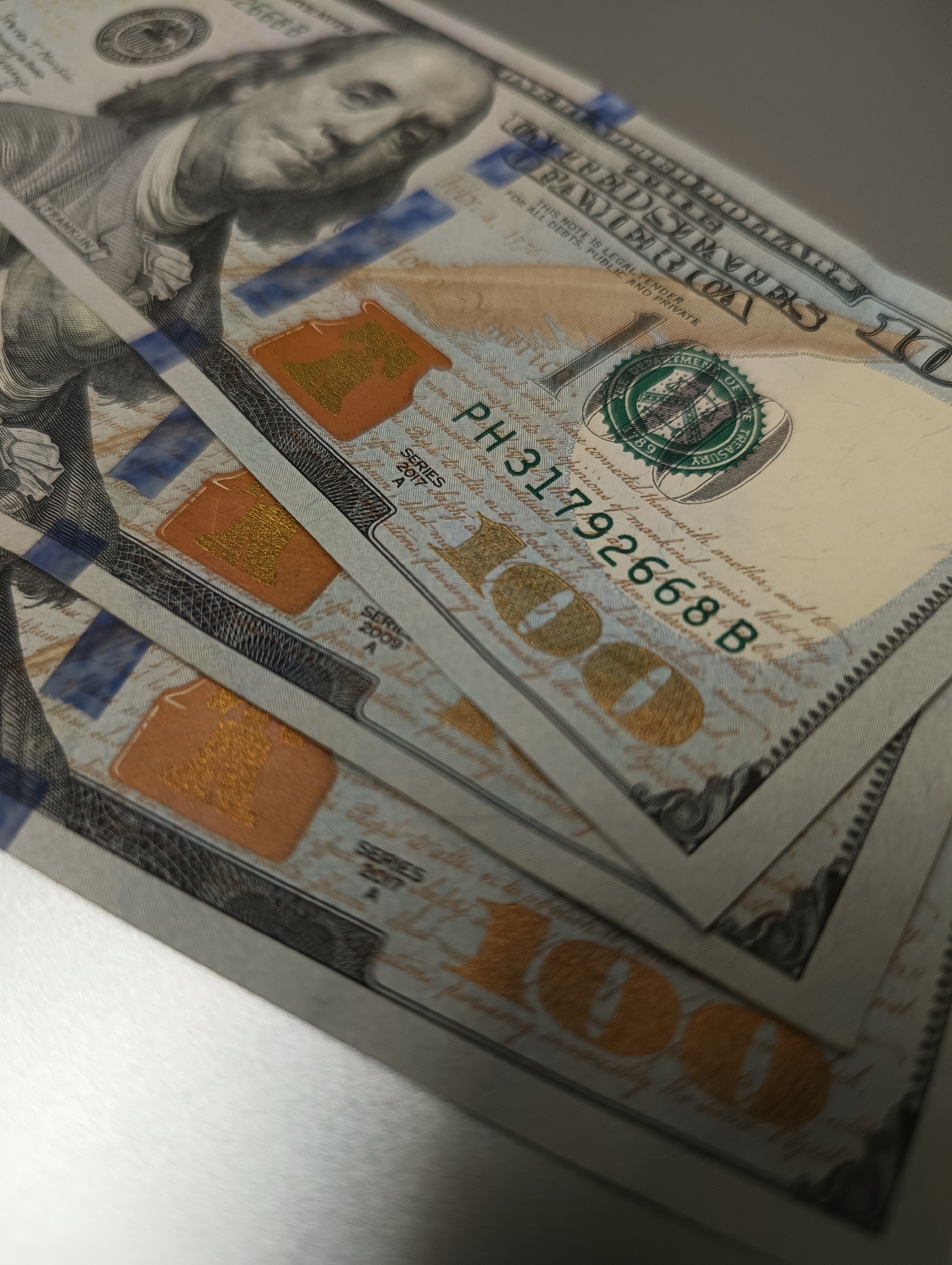 A close-up, overhead shot shows several US one-hundred dollar bills fanned out diagonally across a white surface. The bills are stacked on top of each other, with the top bill displaying Benjamin Franklin's portrait and the serial number "PH31792668B." The bills have various security features, including a blue security thread and a watermark. The lighting casts shadows, obscuring some details of the lower bills. The environment is a plain, white surface, suggesting an indoor setting with no discernible city or location.