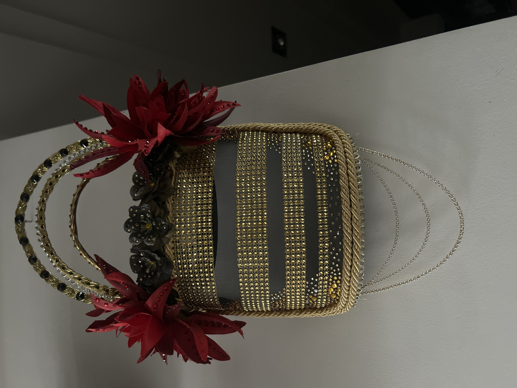 The image shows a decorative handbag with red floral accents placed against a white wall. The handbag features vertical stripes of gray fabric and gold embellishments. It has a twisted gold rope trim and multiple silver chain handles. On the left side, two red artificial flowers and dark gray floral details adorn the bag.
