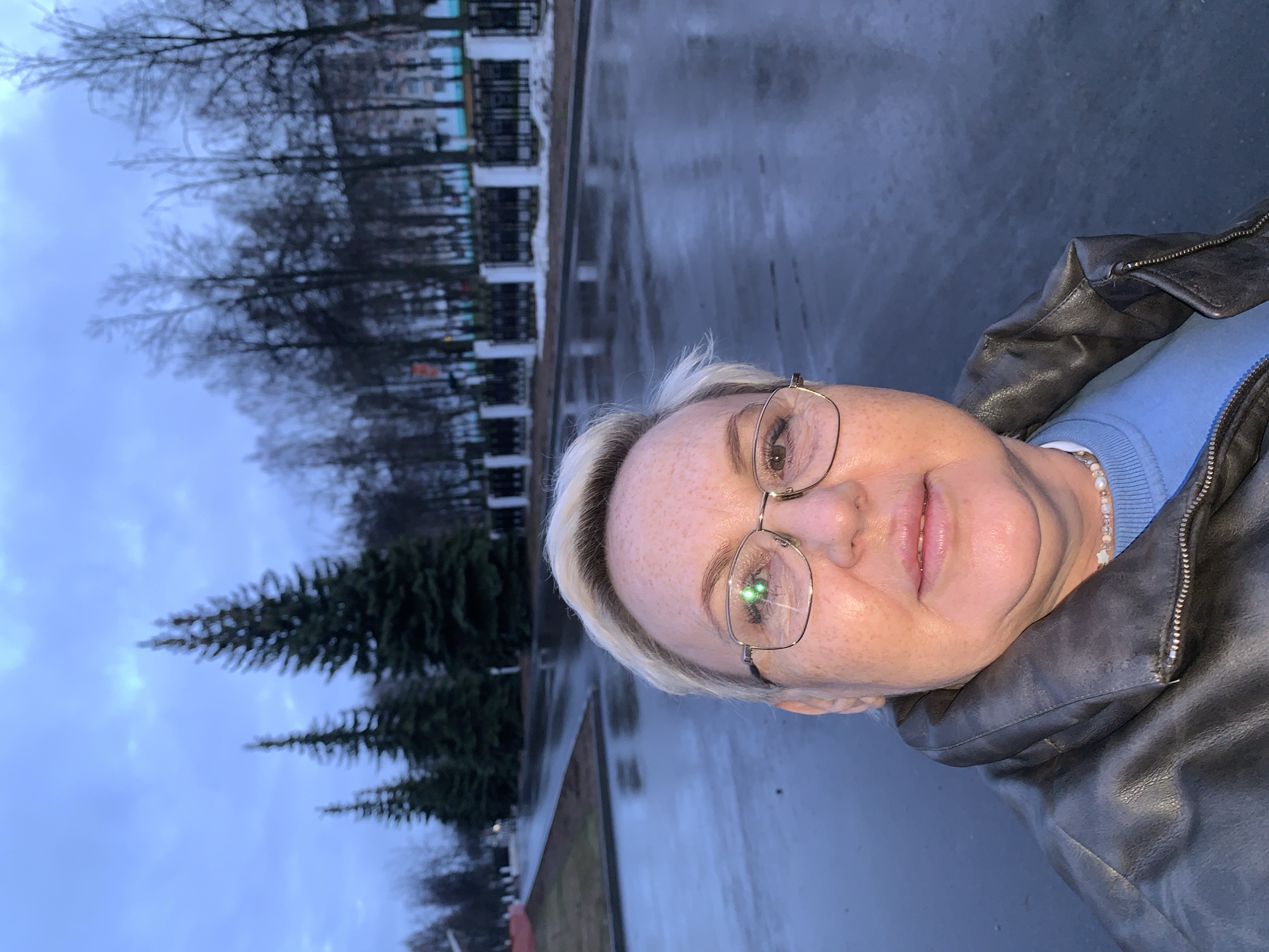 A selfie was taken in what appears to be a park in the city of Chaikovsky, Russia. The person in the image is fair-skinned, with blonde hair and wearing glasses, a light blue sweater, and a dark leather jacket. In the background, bare trees are visible, as well as evergreen trees and a fence. The ground looks wet.