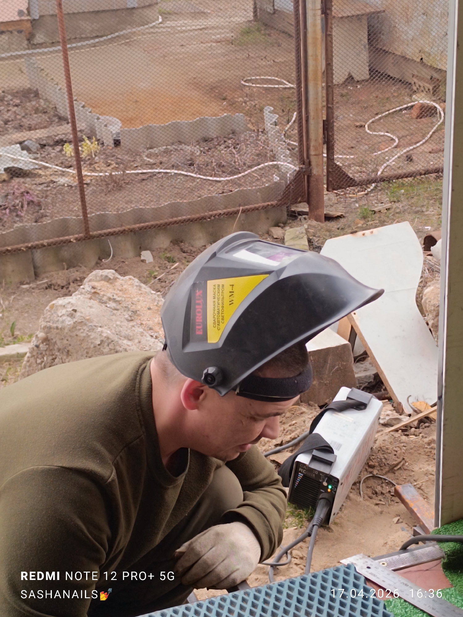 The image depicts a man involved in welding work outdoors in Chaikovsky, Russia. The main subject is a man, seen from the chest up, wearing an olive-green long-sleeved shirt and work gloves. He has a black EUROLUX WM-4 welding helmet, marked with "СВАРОЧНАЯ МАСКА С АВТОМАТИЧЕСКИМ СВЕТОФИЛЬТРОМ" (Welding mask with automatic light filter), pushed back on his head, revealing his concentrated profile. He is crouched down, looking towards his work area.
Next to him on the ground is a silver-colored CAL 50 PECATA welding inverter, which appears to be powered on with green and orange indicator lights visible. The immediate foreground shows a blue, textured surface, possibly a mat or equipment cover, and a metal ruler.
The scene is set in an outdoor, semi-developed environment. The background features dirt ground with sparse vegetation, scattered construction debris such as concrete blocks and pieces of wood, and a rusted metal mesh fence. Beyond the fence, additional structures or parts of a building are faintly visible. The lighting is diffuse, suggesting an overcast day.
Visible text includes watermarks: "REDMI NOTE 12 PRO+ 5G", "SASHANAILS", and the date and time "17.04.2026 16:36". The welding helmet bears "EUROLUX WM-4" and its description, while the welding machine shows "CAL 50 PECATA".