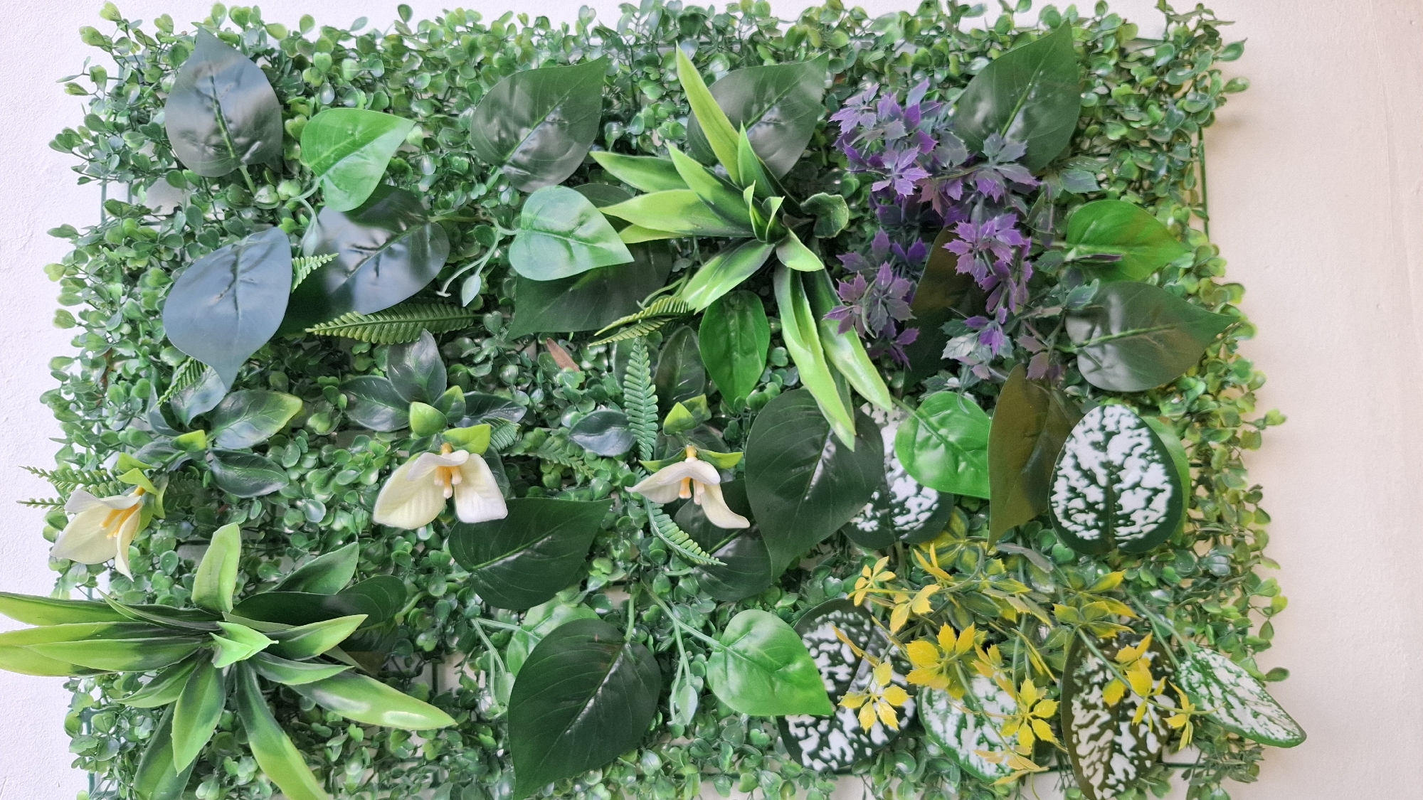 The image shows a square artificial green wall panel with various types of fake plants attached. The plants include several shades of green foliage, small white and yellow flowers, and purple flowers. The panel is mounted against a light beige or off-white wall. The environment suggests an interior space. There are no people visible in the image. There is no information in the image to identify the location as Sofia, Bulgaria.