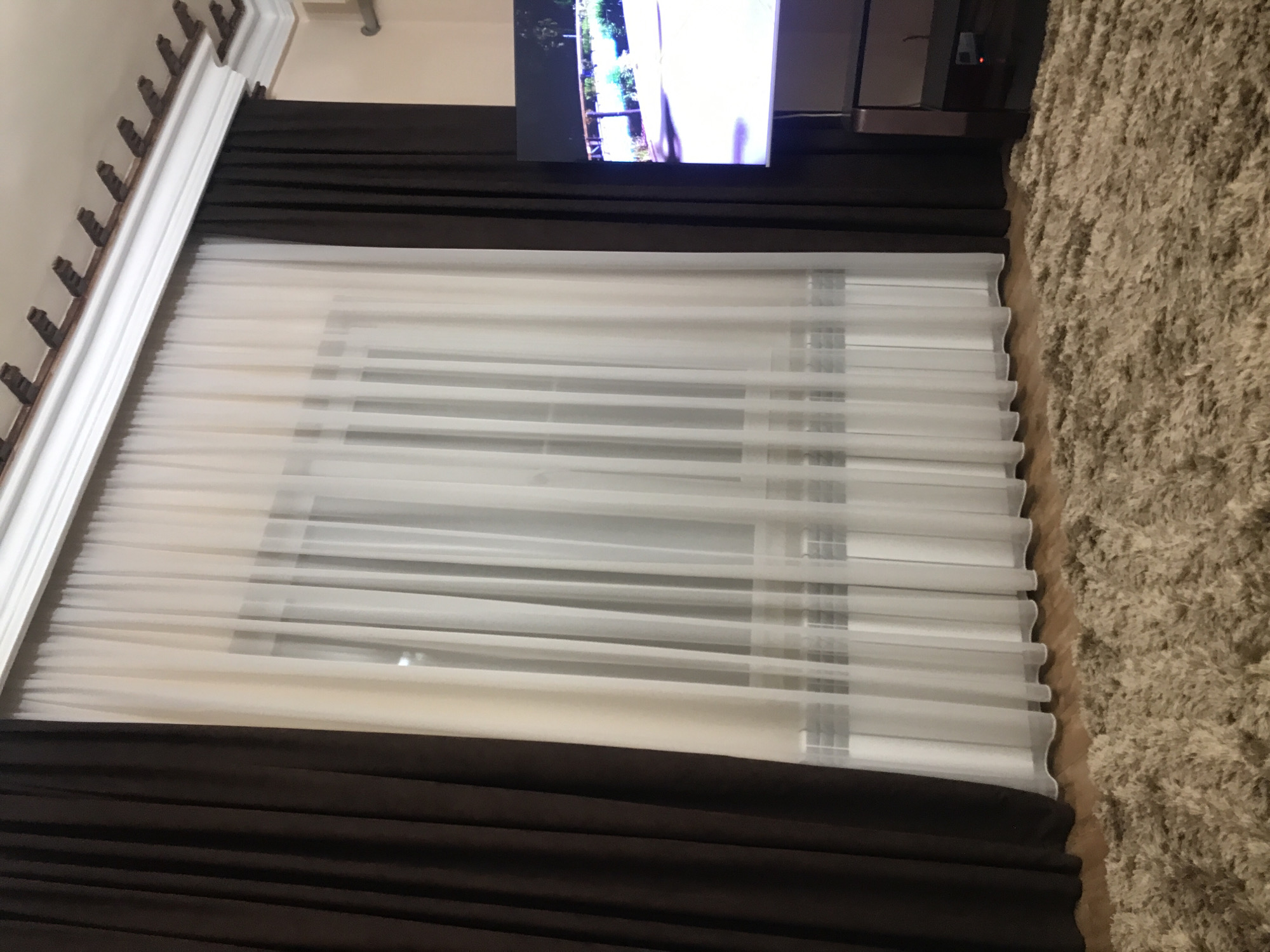The image shows a window covered with white and brown curtains, a television mounted above the window, and a beige shag rug partially visible on the right side of the frame. The wall near the window is adorned with decorative white molding. The window appears to have white blinds. The location is potentially a residential or hotel room. There is no information available to confirm if this image was taken in Yerevan, Armenia.